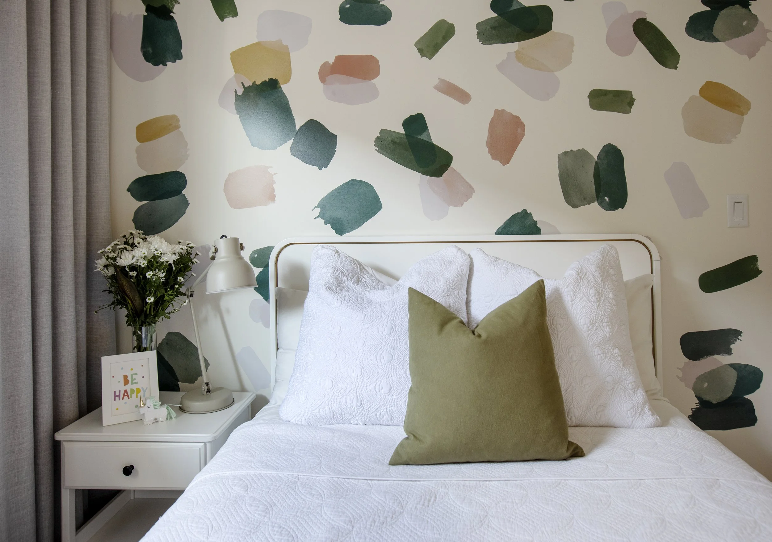 A bedroom with a white bed, green and white pillows, a white nightstand with a lamp, a bouquet of white flowers, and a colorful wall mural featuring abstract shapes in green, yellow, pink, and beige.