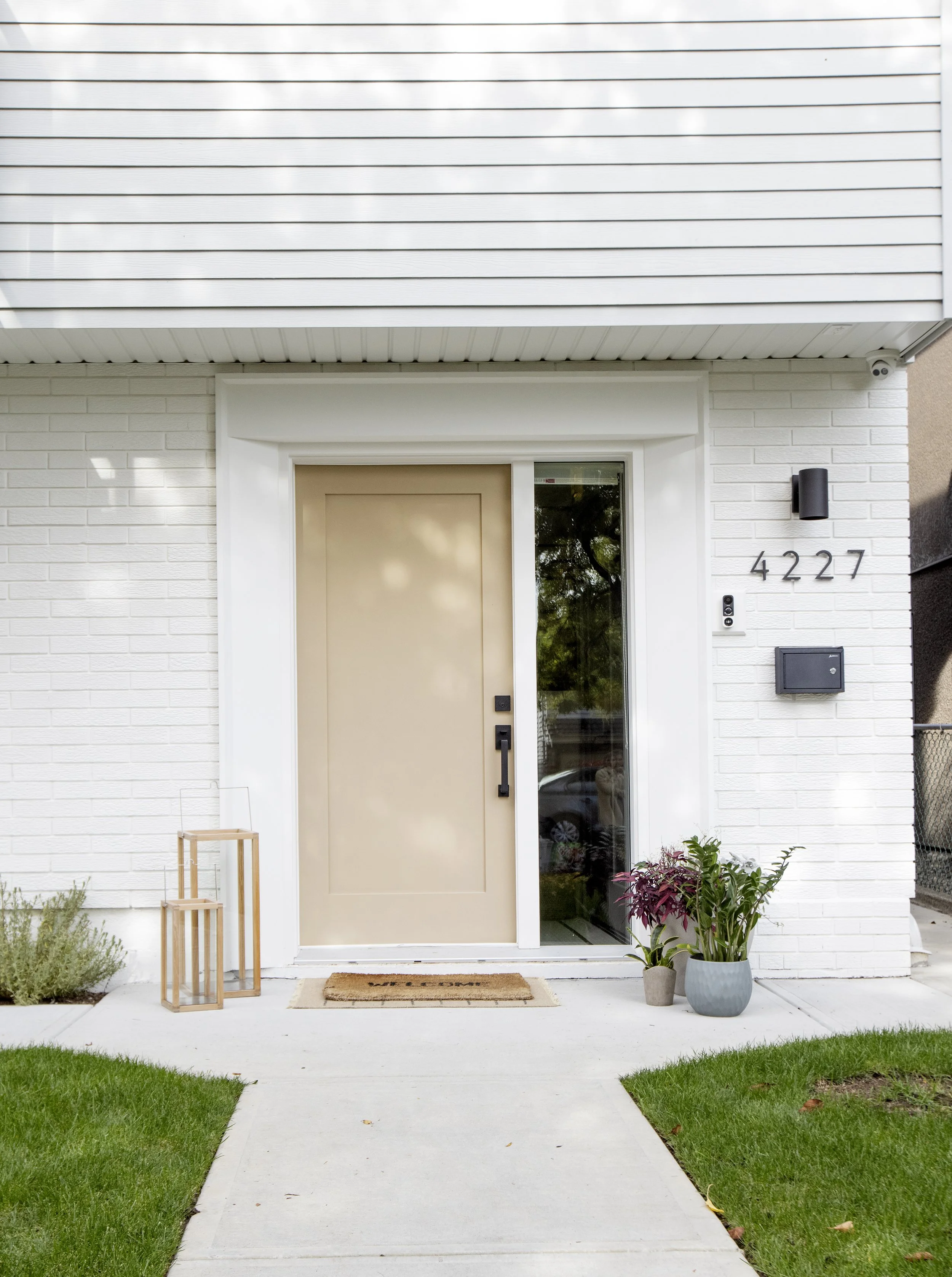 Front door of a modern white brick house with a beige door. There are two potted plants, one on each side of the entrance, and a small wooden table on the left. The house number 4227 is displayed on the right side of the door. There is a security cam