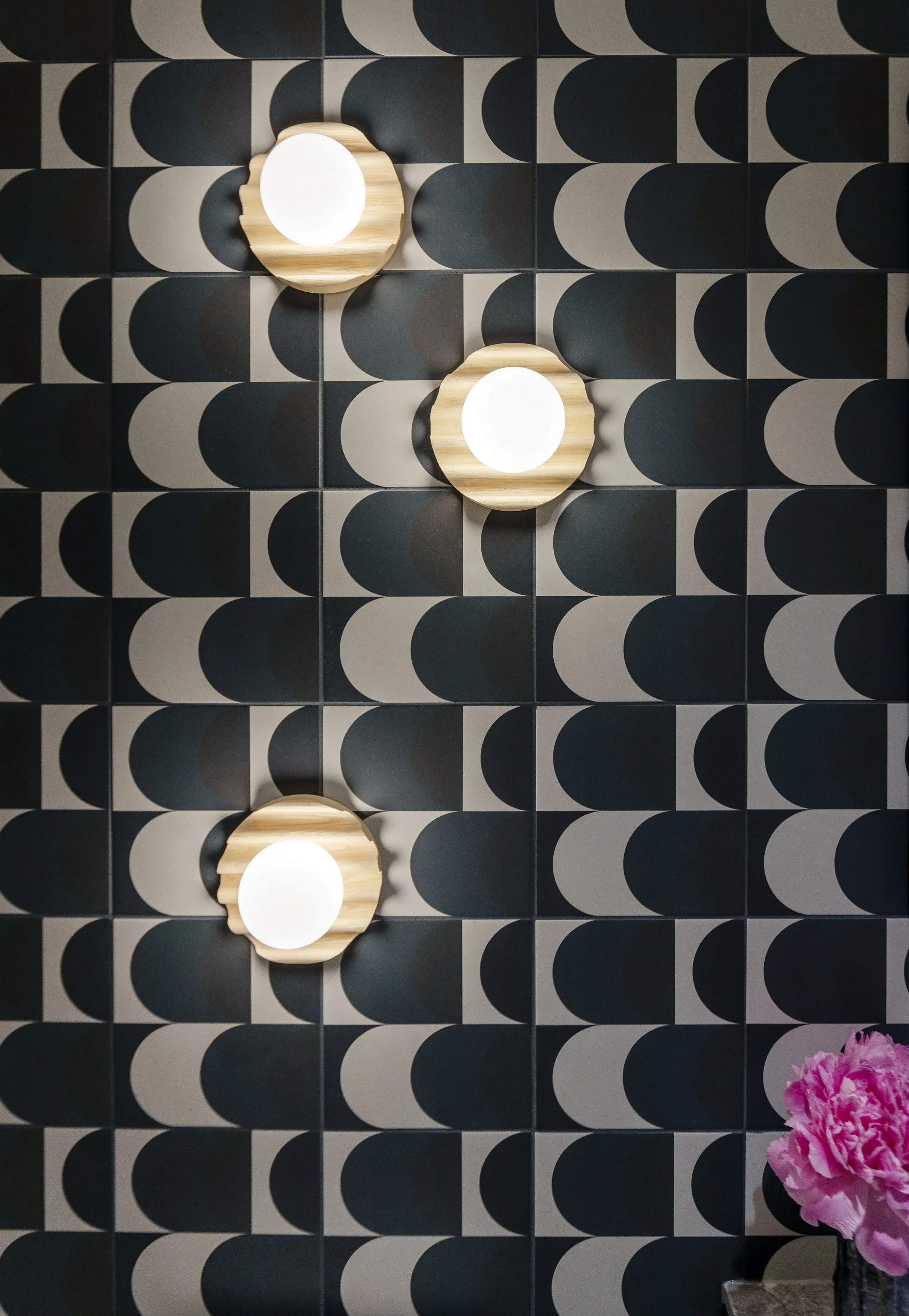 Decorative wall with a black and gray geometric pattern, featuring three round wall lights with wooden surrounds and white diffusers, and a pink flower arrangement in a black vase in the bottom right corner.