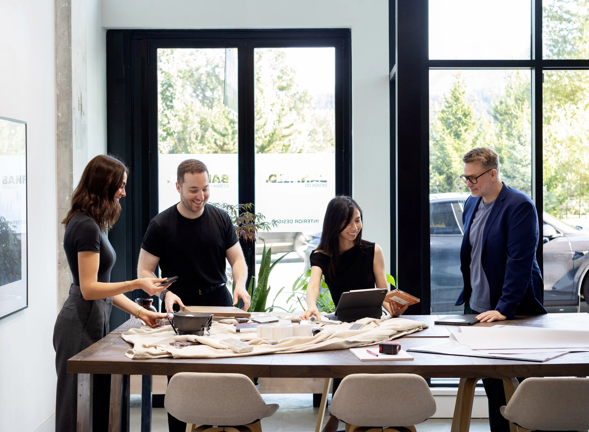 People gathered around a table in a modern office with large windows, looking at various items and working on a project.