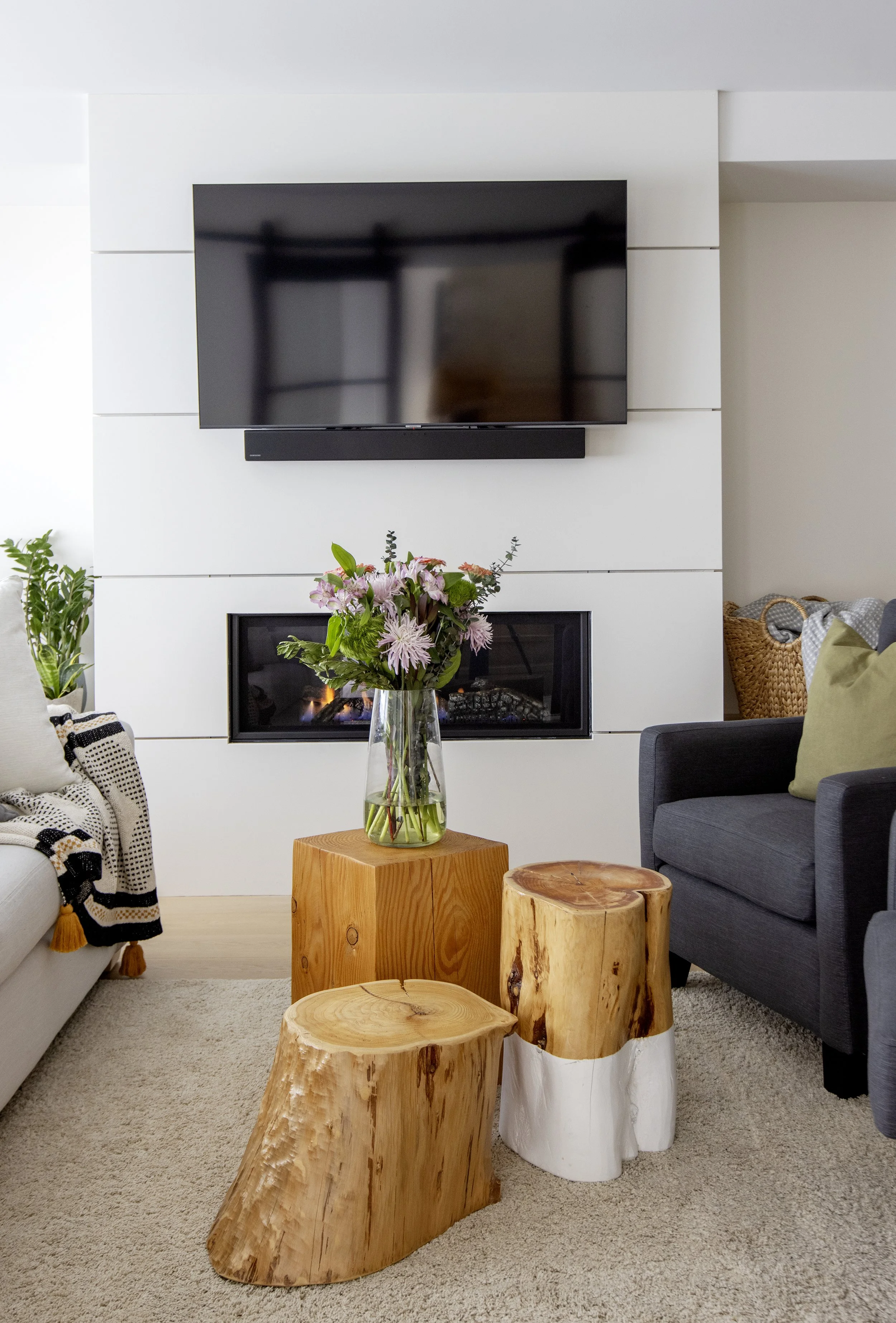 Living room with a wall-mounted flat-screen TV, a fireplace with a floral arrangement on a glass vase, and wood and fabric furniture including a gray armchair, a white sofa, and various wood stools on a beige carpet.