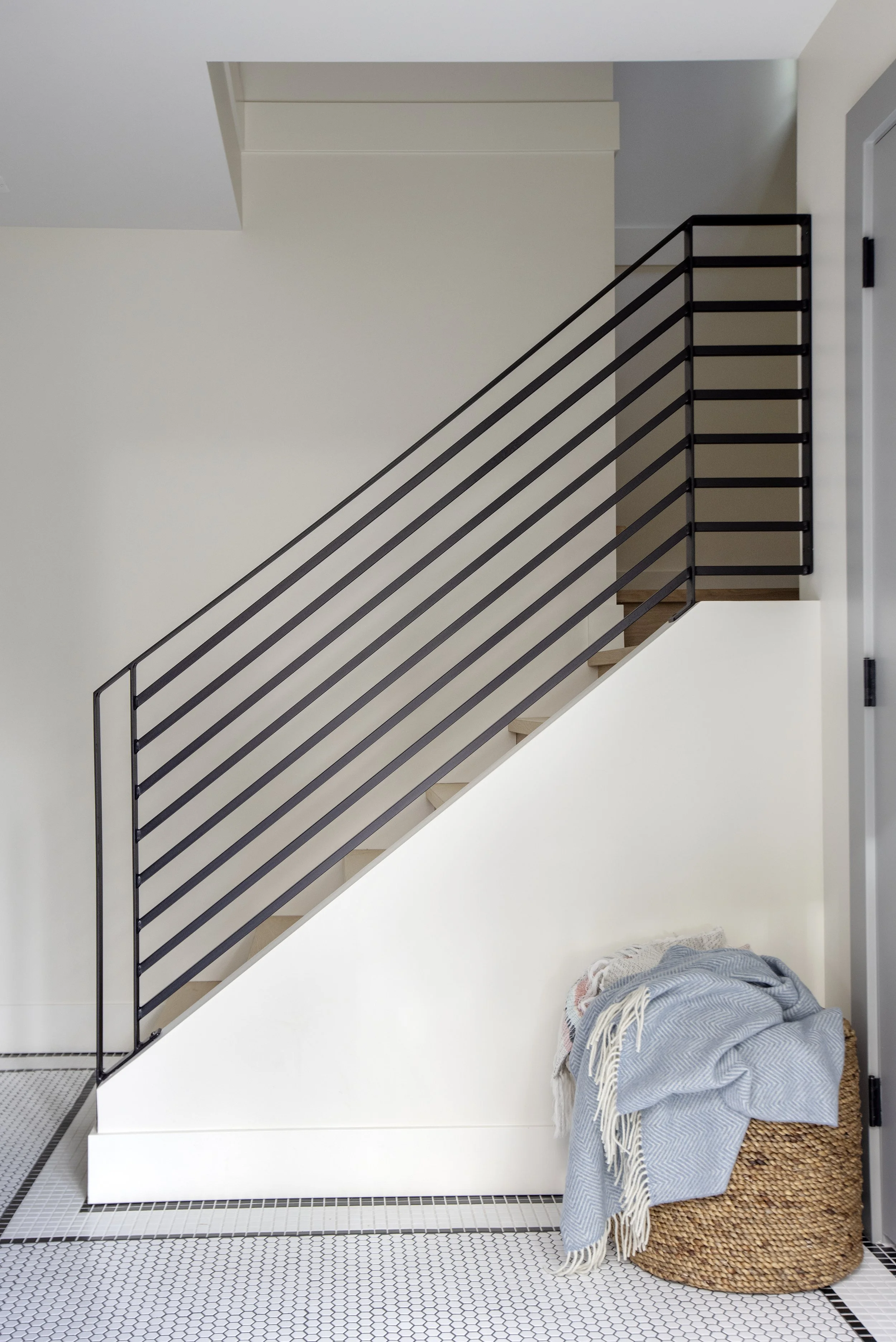 Interior view of a modern staircase with black metal railing, white walls, tiled black and white hexagonal floor, and a bundle of blankets on a woven basket underneath the stairs.