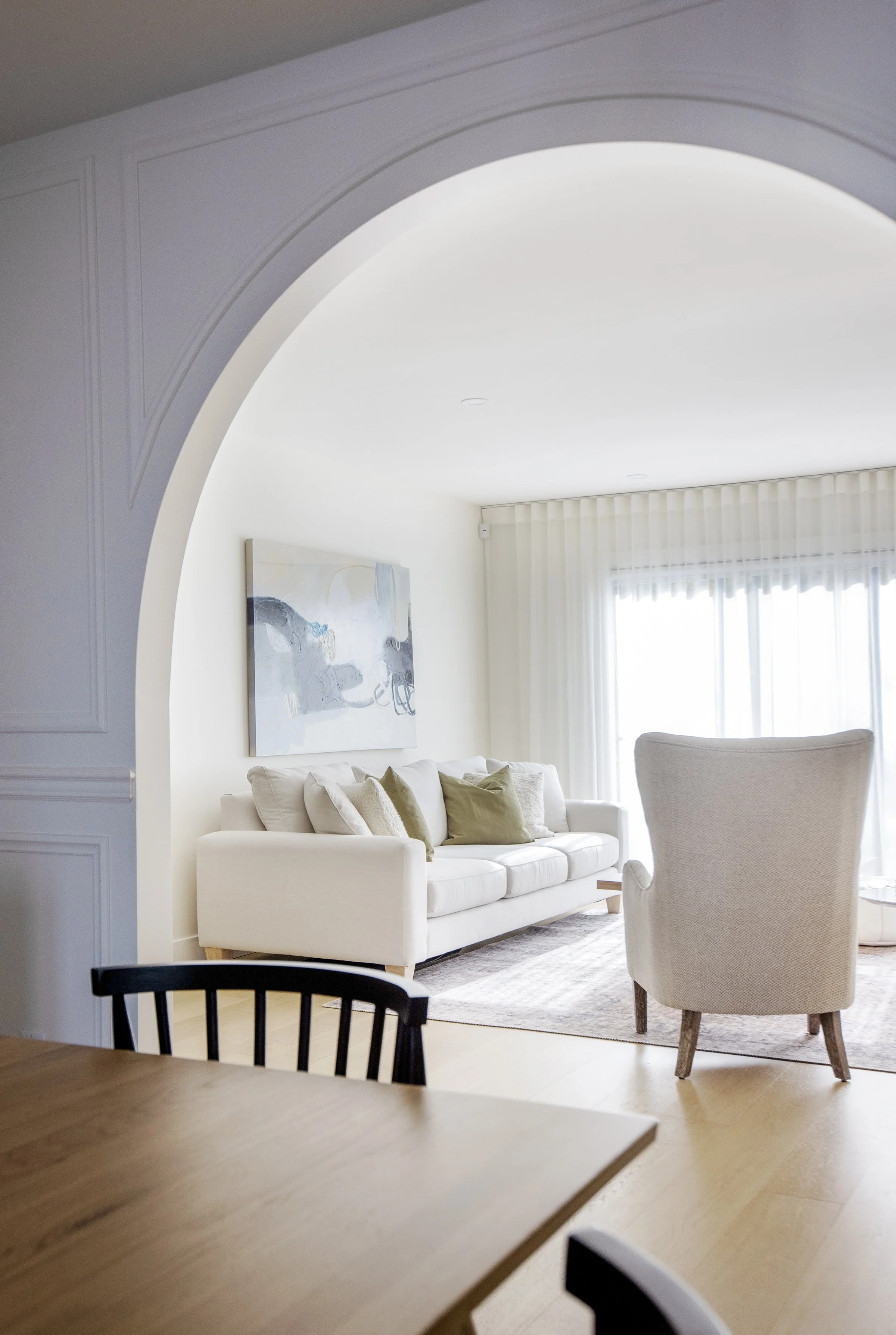 A bright living room with a white sofa, olive green pillows, a large abstract painting, a high-backed armchair, and sheer curtains over a window, viewed from the dining area with a wooden table and black chairs.