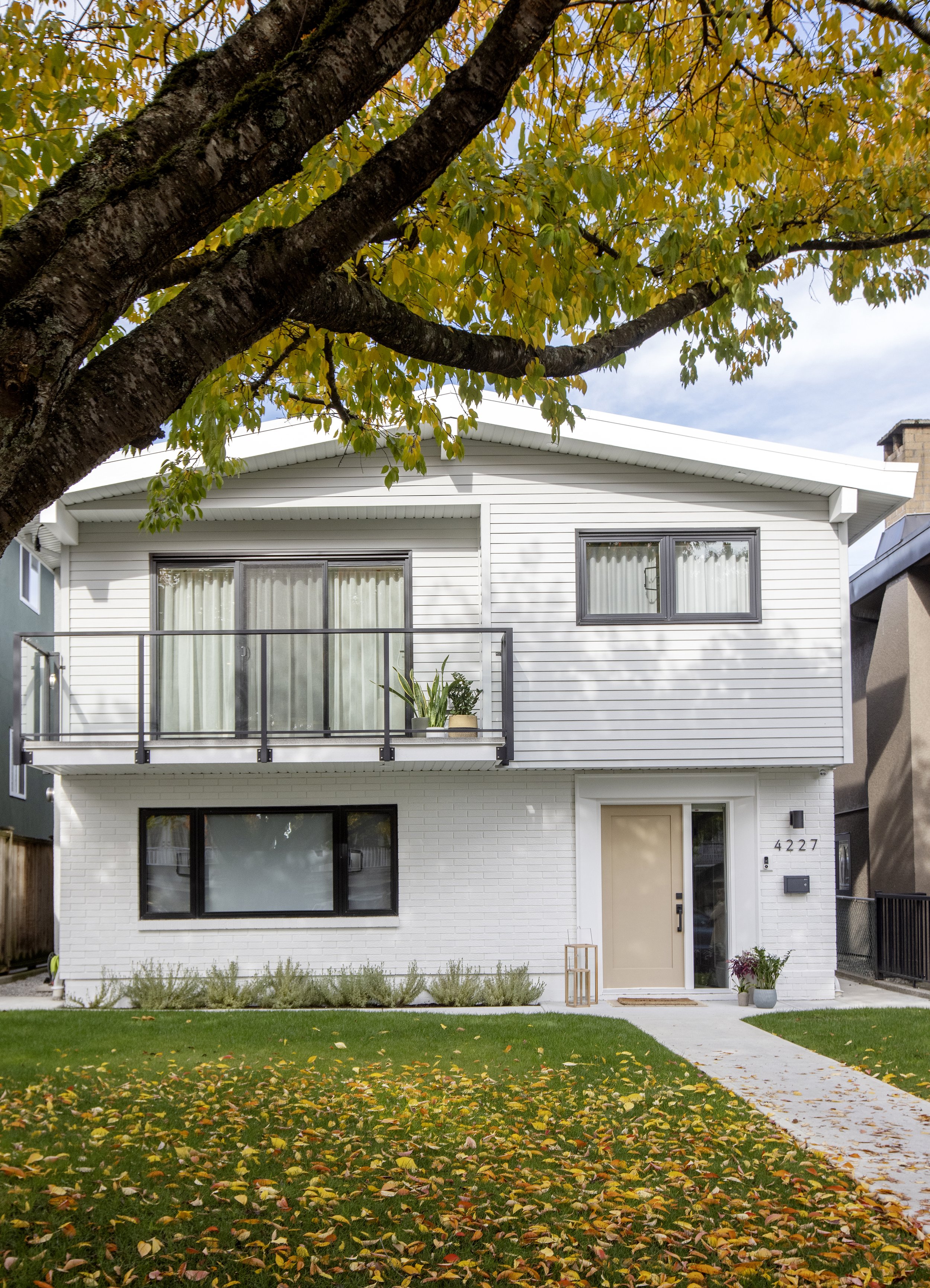 A white multi-story house with large windows, a balcony with glass railing, and a front door. The house number is 4227. There are plants near the entrance and a garden with grass and fallen yellow leaves. A sidewalk runs in front of the house, and a 
