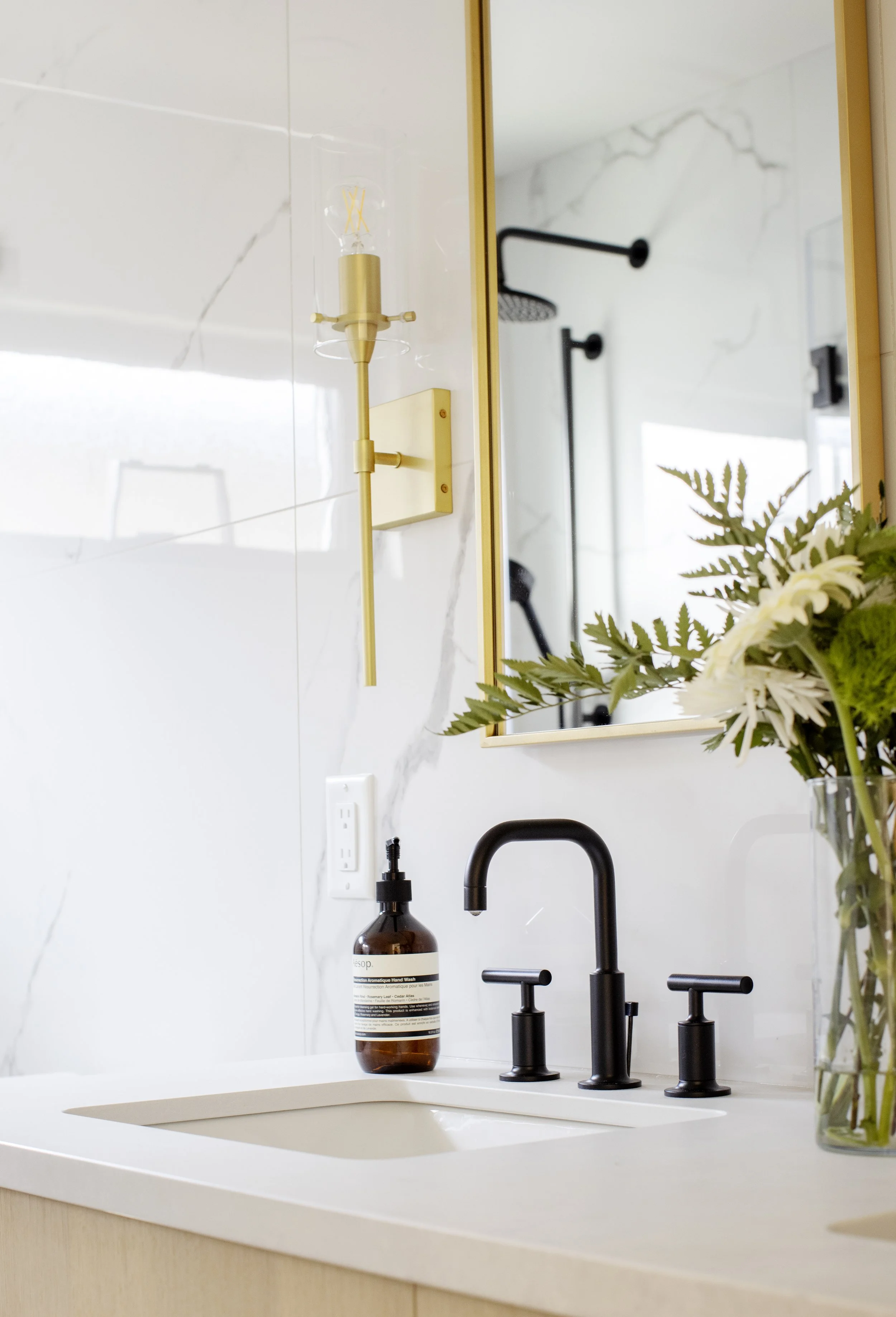 Close-up view of a modern bathroom sink with a black faucet, a brown hand wash soap dispenser, a gold wall-mounted light fixture, a large mirror, a green potted plant in a clear glass vase, and a white marble backsplash.