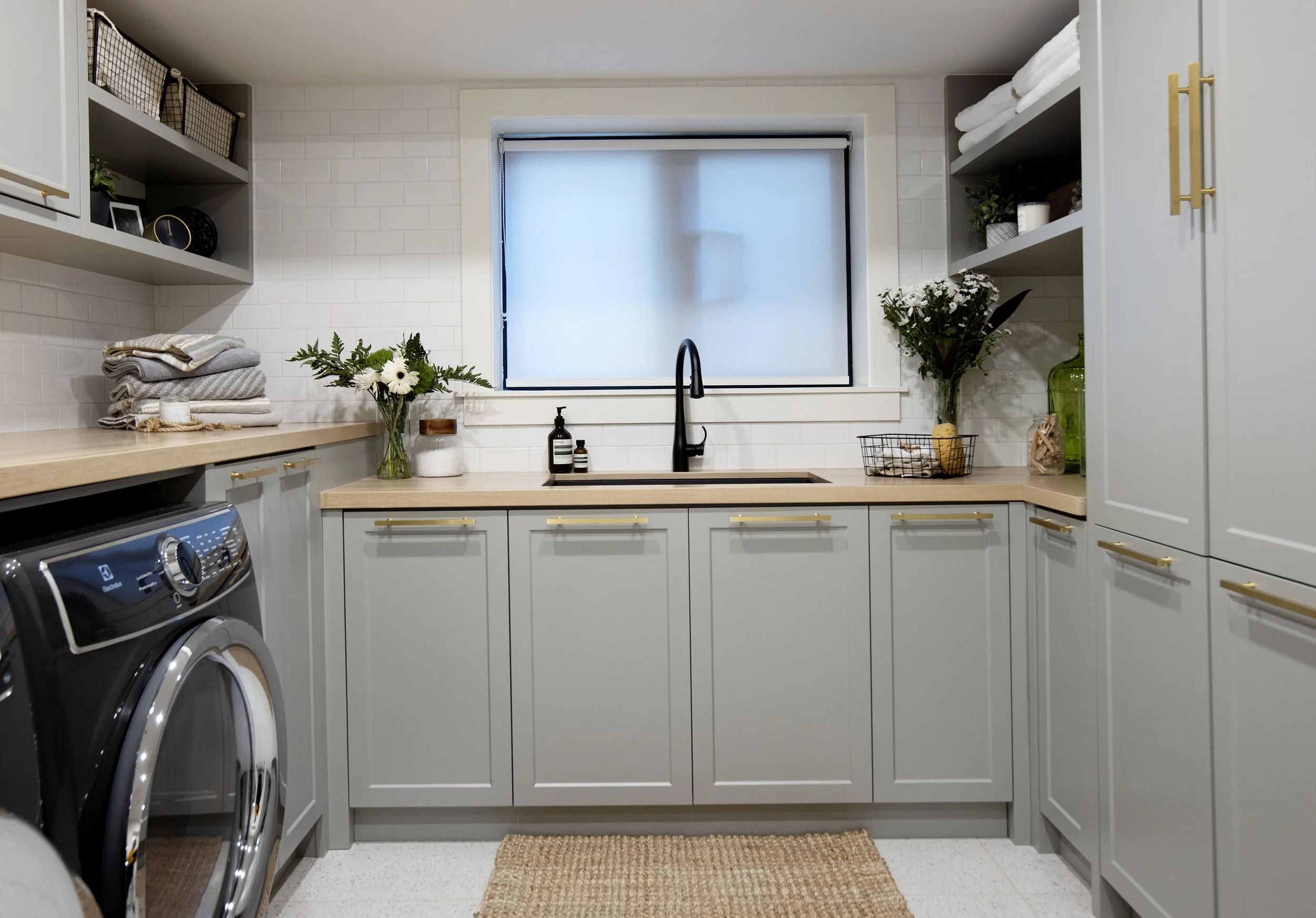 A washer and dryer in a laundry room with white cabinets, open shelving with towels and decorative items, a window with frosted glass, and a tan rug on the floor.