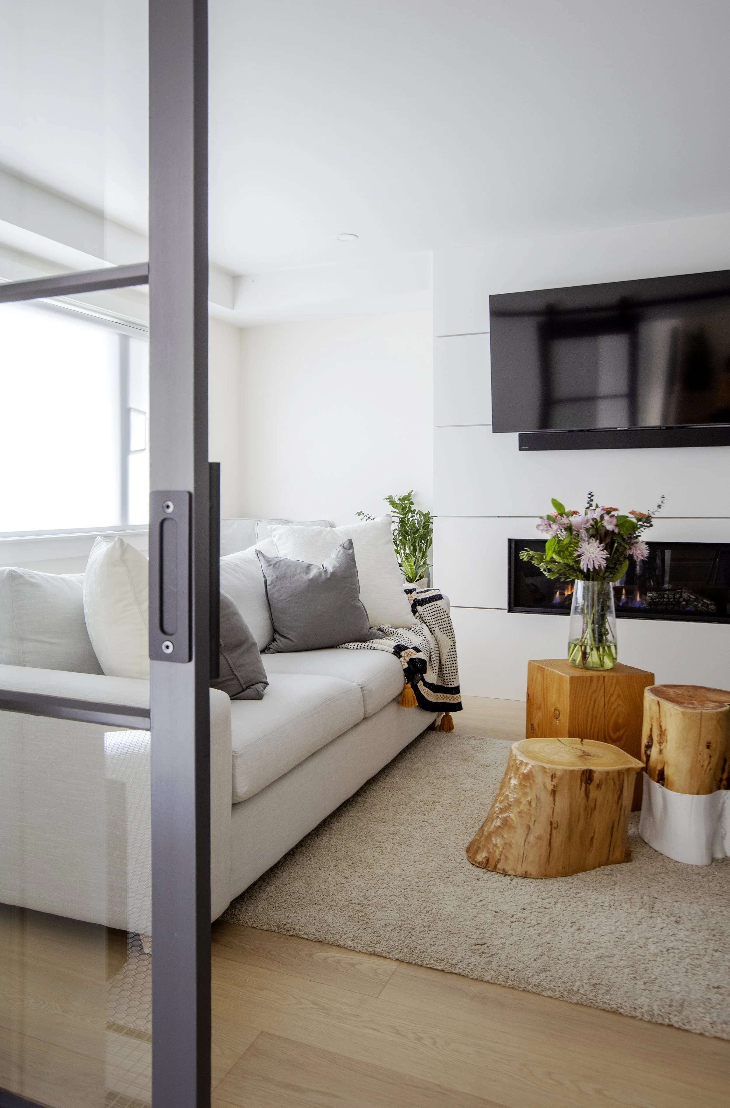 A living room with a white sofa, gray and white pillows, a beige area rug, and a stone fireplace with flowers on the mantel. There are wooden stump side tables and green plants, and a black wall-mounted TV on white wall panels.