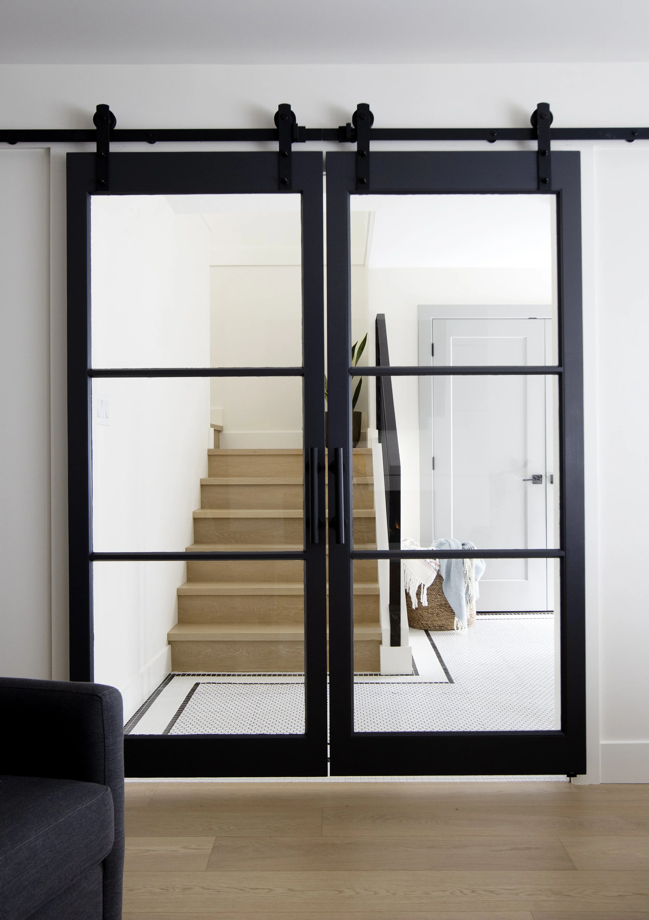 Interior view of a house entrance with black barn door-style sliding doors, wooden stairs leading up, and a laundry basket with cloth near a white door, with a black and white patterned tile entryway and light wood flooring.