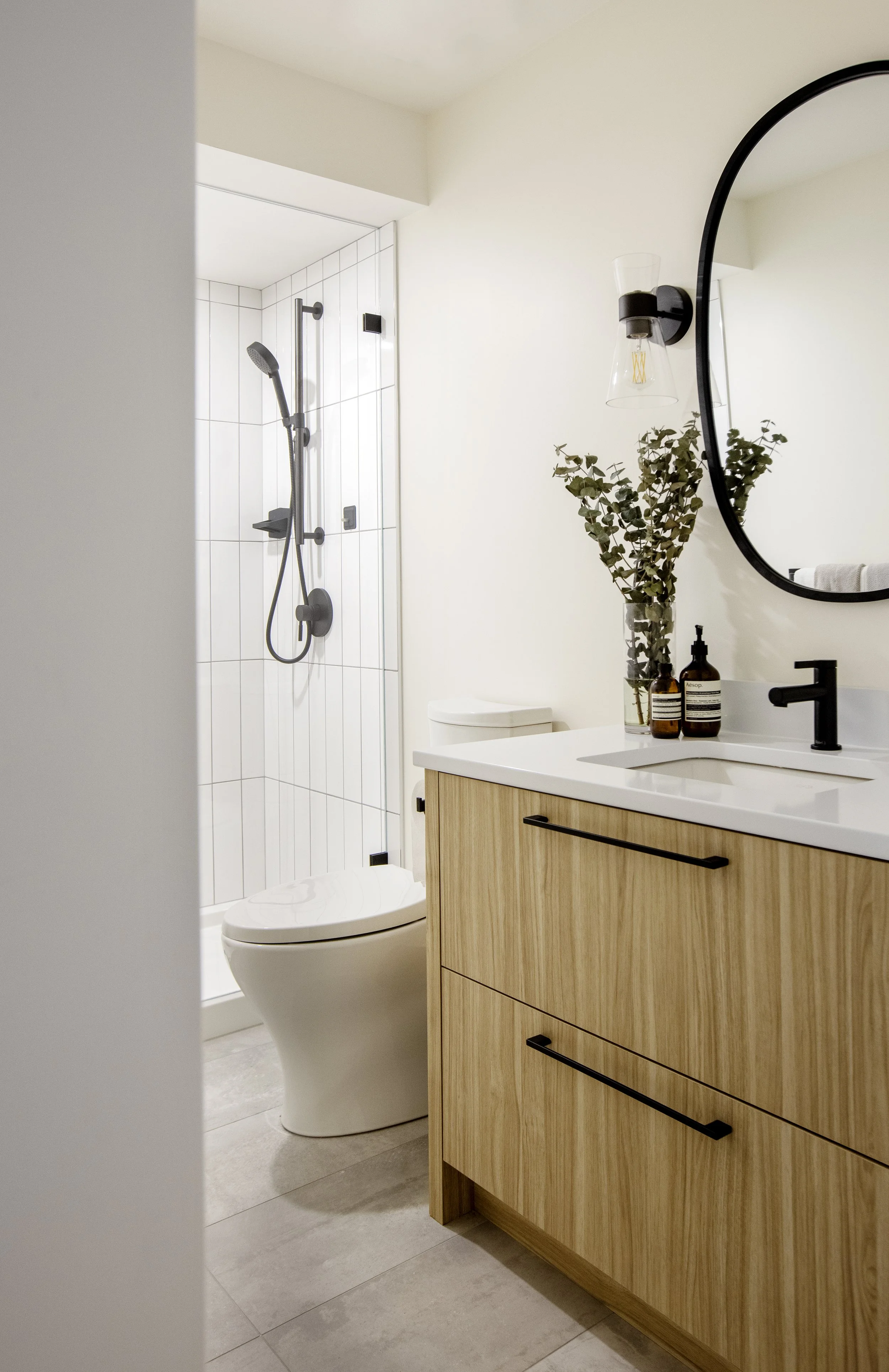 Bathroom with a shower, toilet, and wooden vanity with black handles and a large mirror, decorated with a vase of green foliage and soap bottles, illuminated by natural light.
