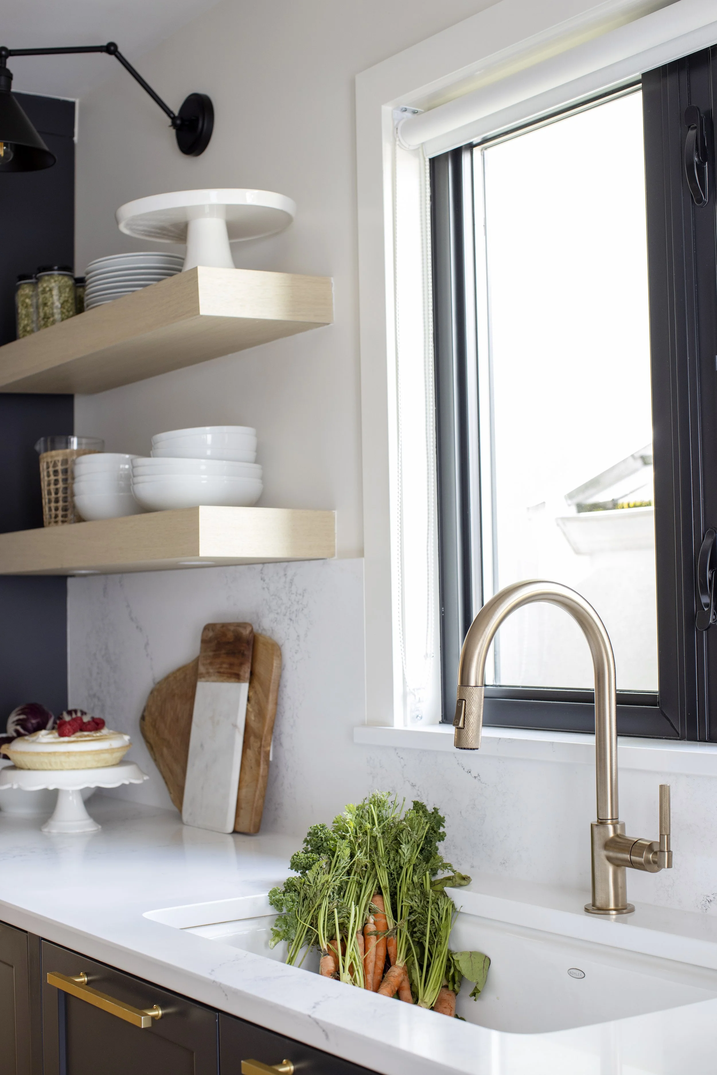 A modern kitchen with a white marble countertop, dark cabinetry with gold handles, and a window with black trim. Fresh carrots with green tops are in the sink, a cake with berries is on a stand, and various dishes and jars are on floating shelves. A 