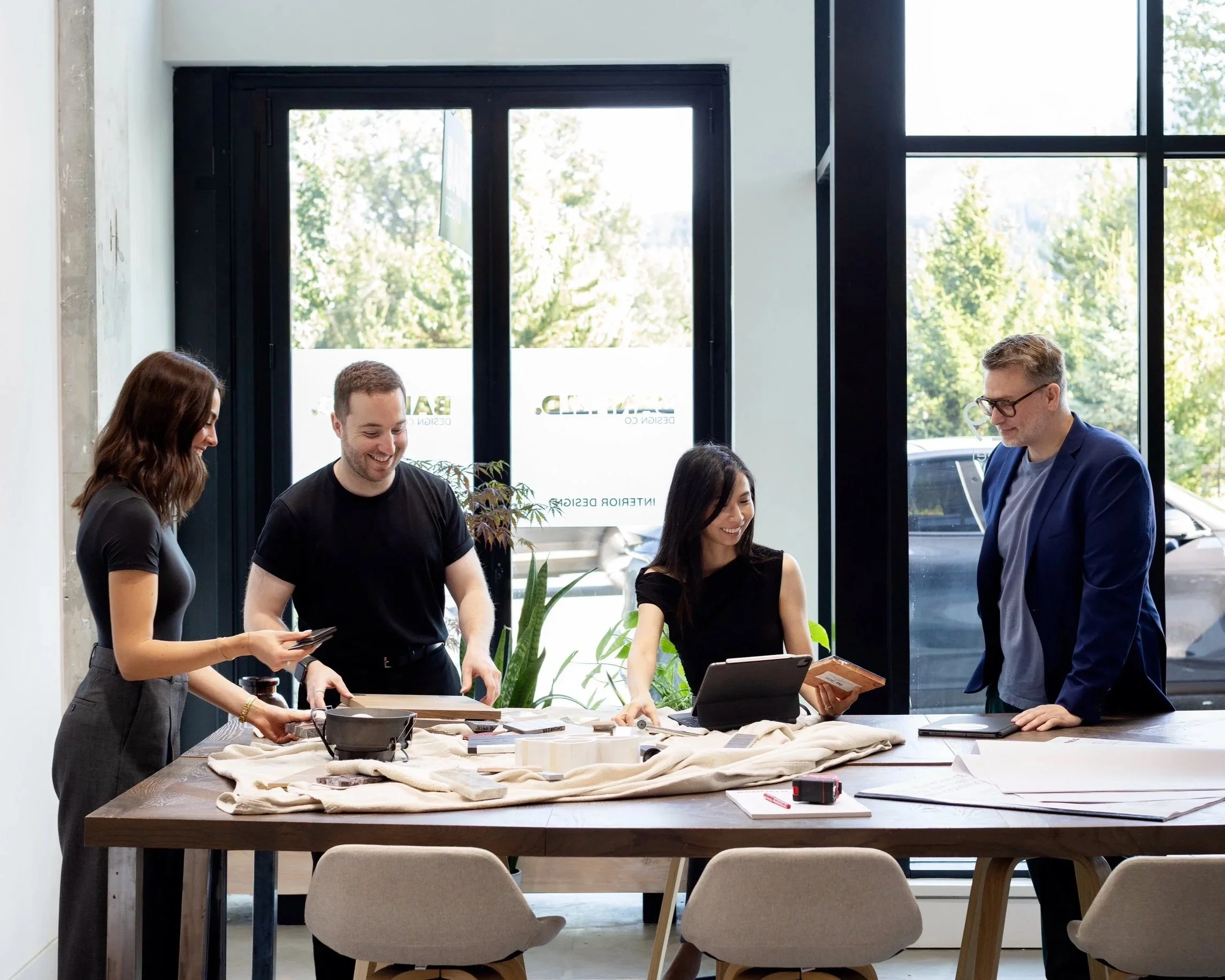 A group of five people gathered around a table in a bright, modern space with large windows, engaged in a discussion or collaborative project. The table is covered with various materials and devices, including a laptop and papers.