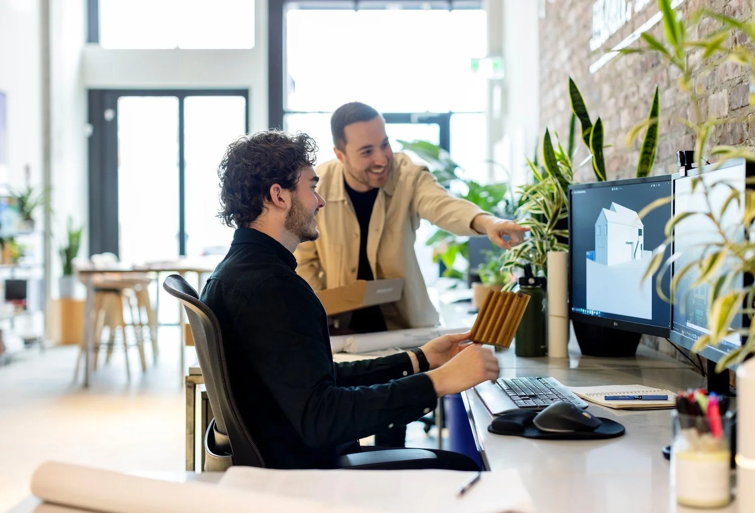 Two men collaborating in a bright, modern office with greenery, working on a 3D architectural design on computer screens.