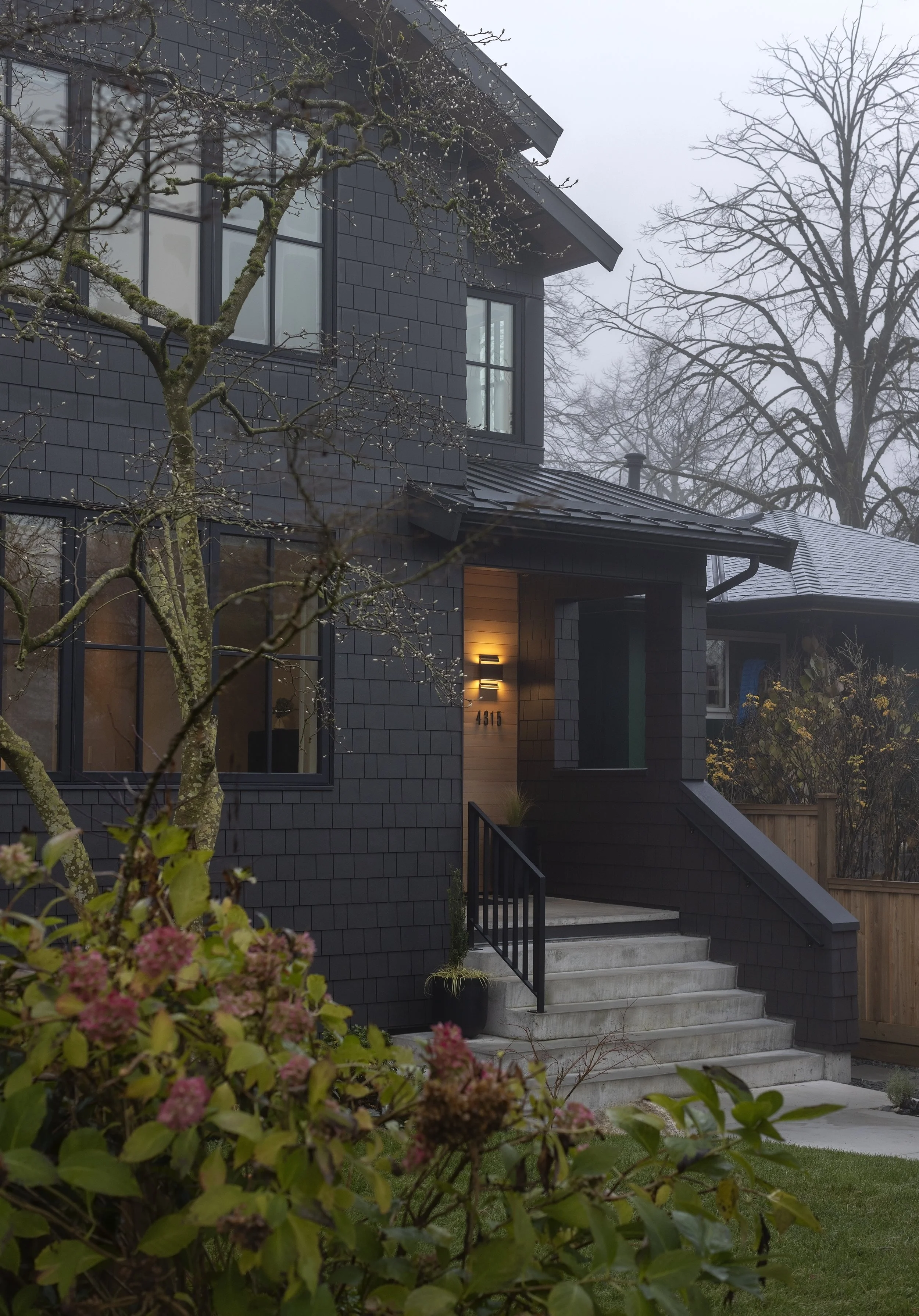 A modern two-story house with dark exterior siding and large windows, surrounded by trees and a landscaped yard with pink flowering bushes and green lawn. The front entrance has concrete stairs, black railing, and a porch light.