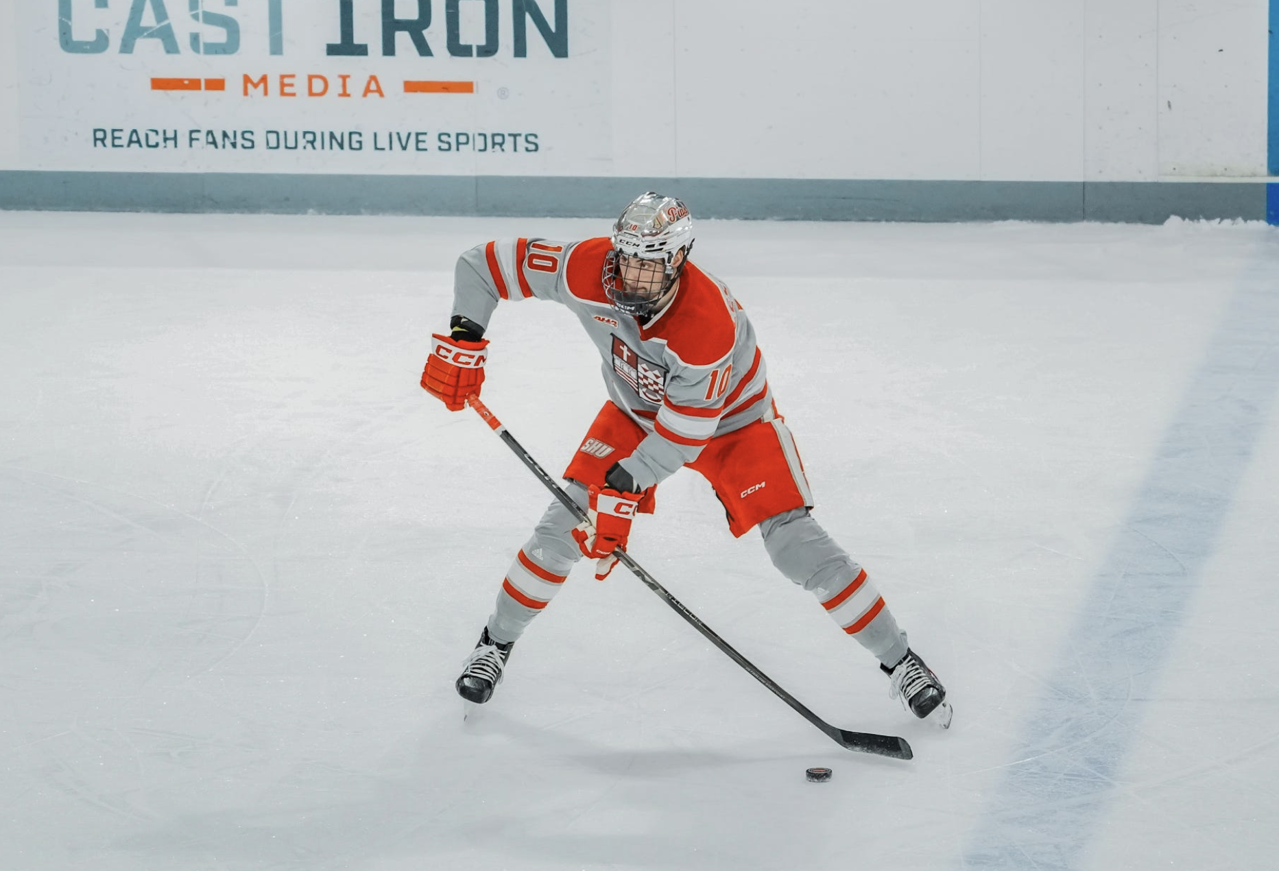 An ice hockey player in a gray and orange uniform is skating and handling a hockey puck on the ice, with a large banner in the background reading 'Cast Iron Media' and 'Reach Fans During Live Sports'.