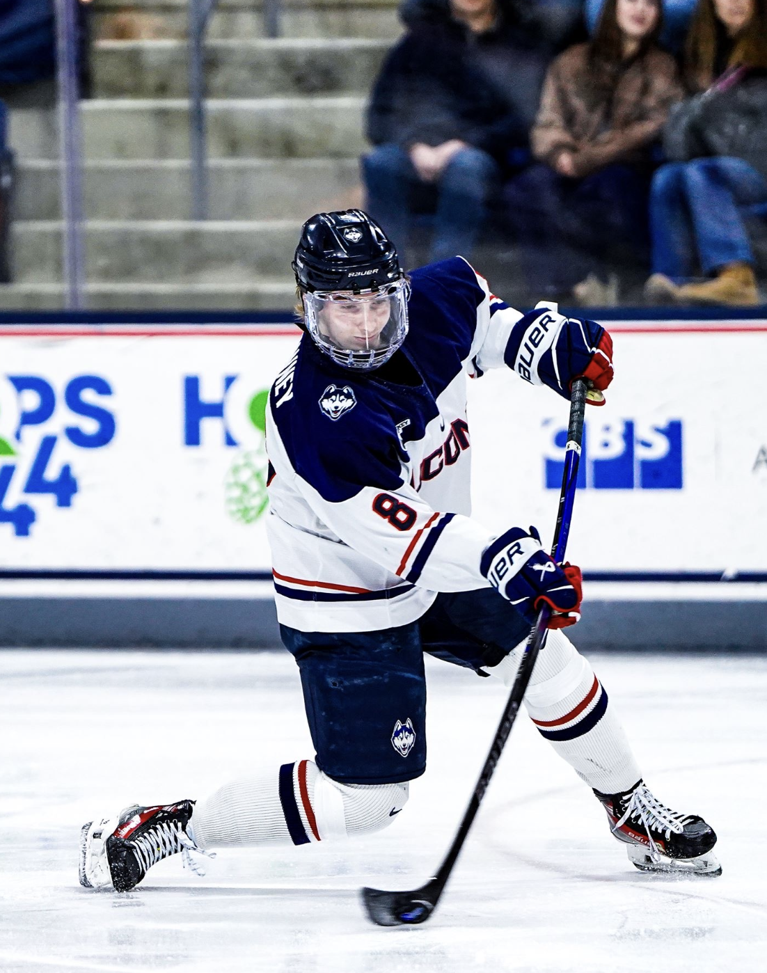 Ice hockey player in a white and blue uniform with the number 8, wearing a helmet, skating on ice with a hockey stick, in an arena with spectators in the background.
