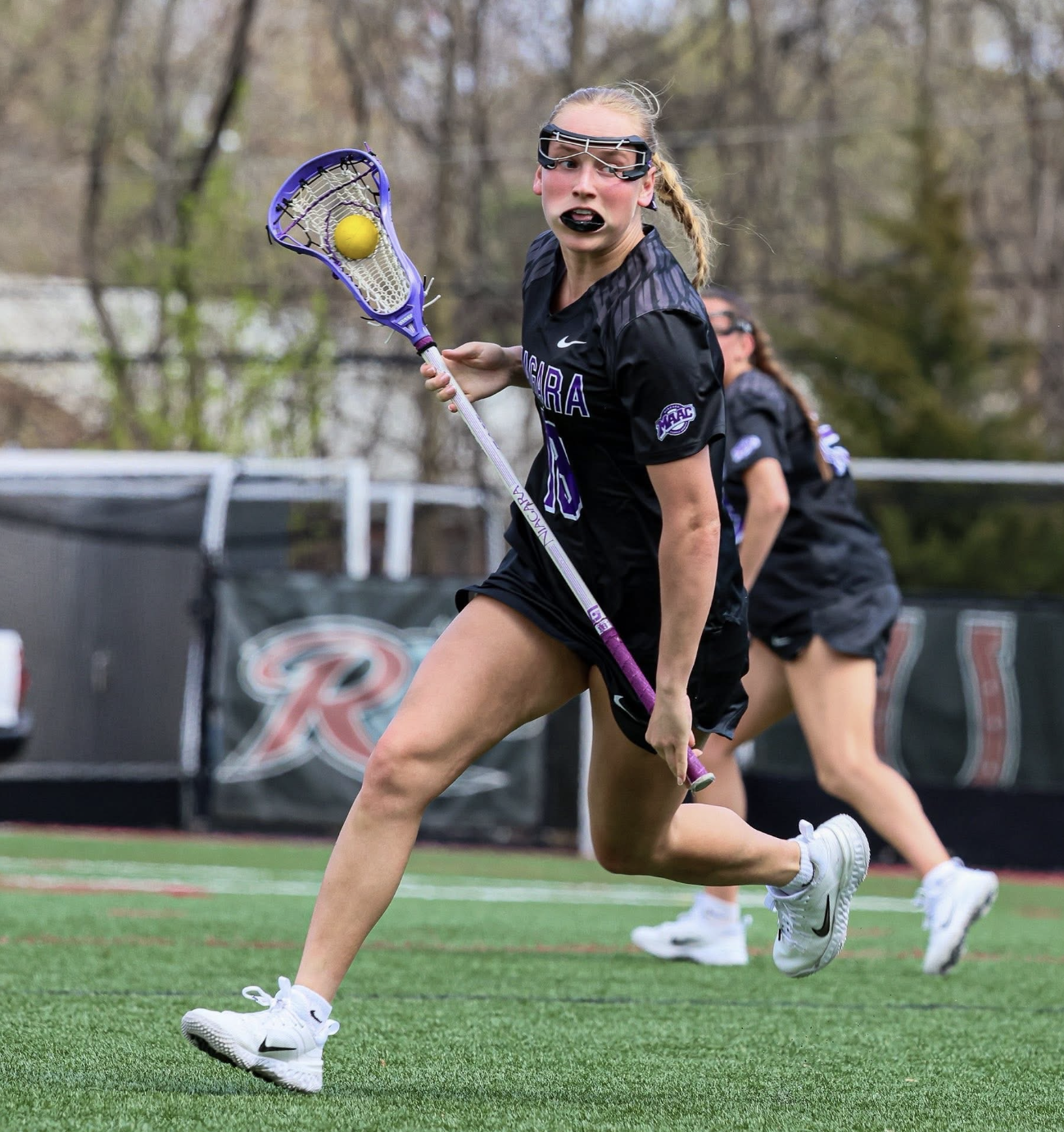 A female lacrosse player in black uniform with purple accents running on a field with a yellow lacrosse ball in her stick's pocket, wearing white Nike shoes, black goggles, and black lipstick, with her hair tied back, and two other players in the background also running.