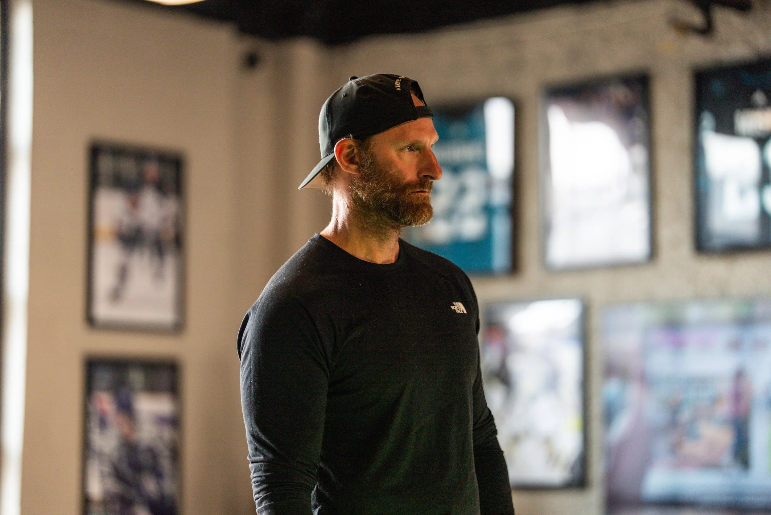 A man with a beard wearing a black cap backwards and a black long-sleeve shirt, standing indoors with posters on the wall behind him.