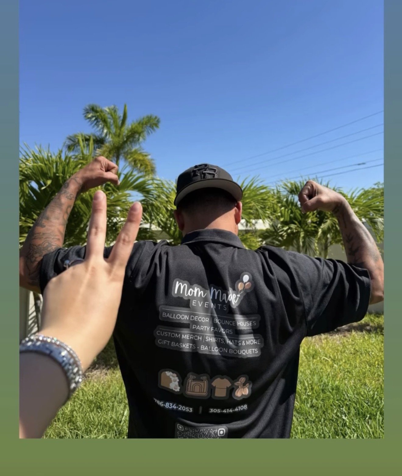 A person wearing a black shirt and a cap is flexing their biceps in front of palm trees and a clear blue sky, with a hand showing a peace sign in the foreground.