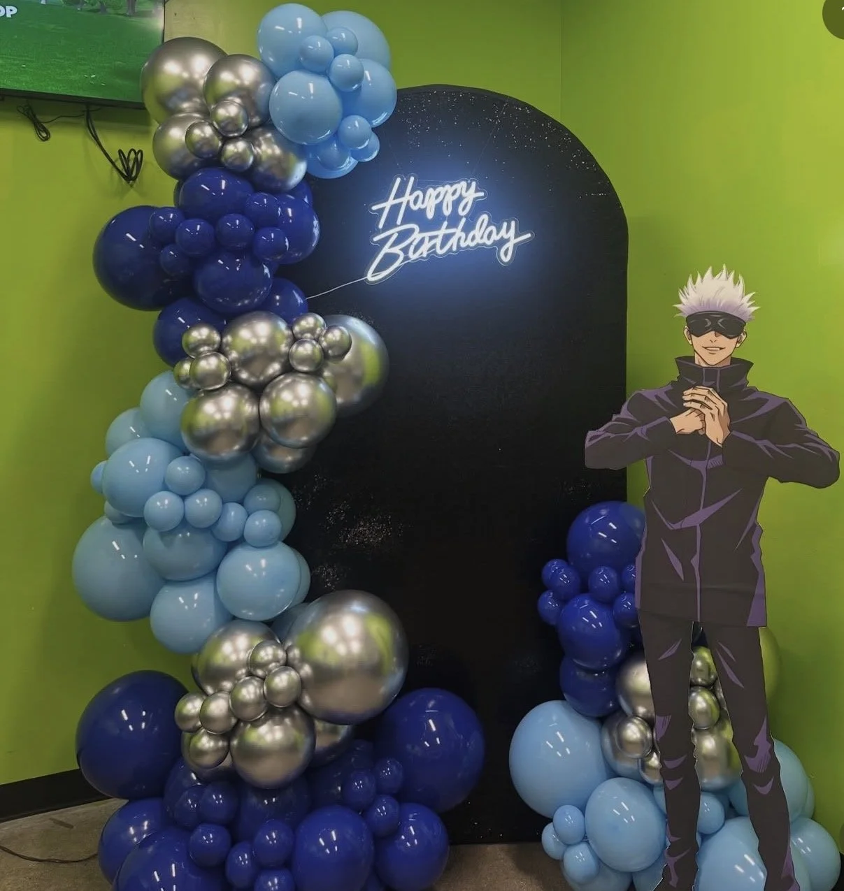 Balloon arrangement with shades of blue, silver, and black balloons, a black backdrop with a neon sign that says "Happy Birthday," and a life-sized cardboard cutout of a character with spiky white hair, sunglasses, and a dark jacket, standing in front of a green wall.