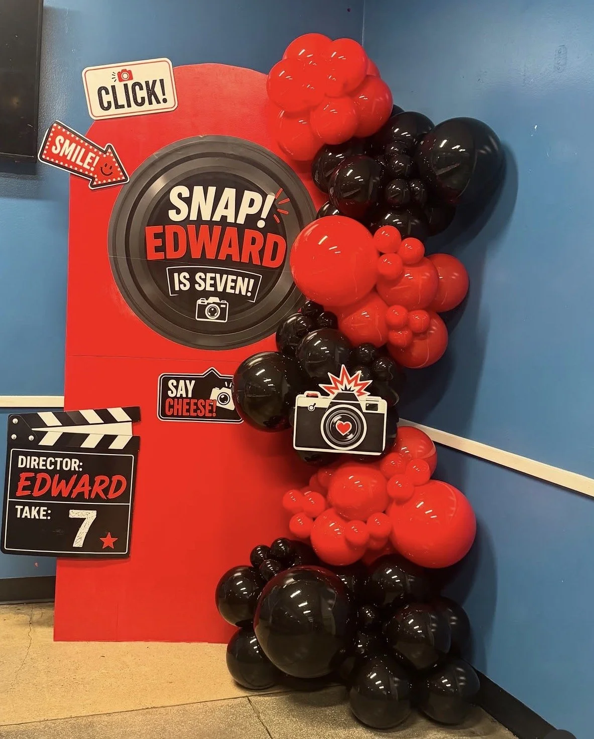 Red and black balloons arranged on a wall for a birthday celebration. A decorative display features a sign with the name 'Edward' and the number seven, along with various playful signs like 'Click!', 'Smile!', 'Say Cheese!', and a film clapboard.