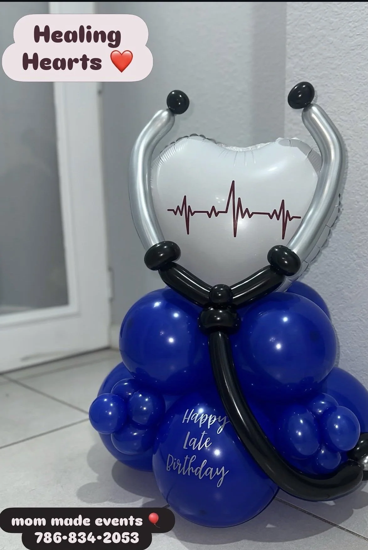 Balloon decoration in the shape of a stethoscope with a heart rate line on a white balloon, surrounded by blue balloons with the message "Happy late Birthday." A label at the bottom indicates "mom made events" with a phone number.