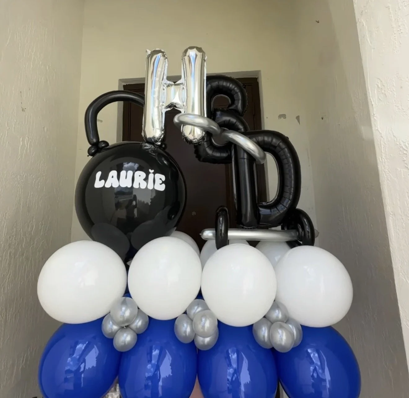 Balloon arrangement with large blue, white, and silver balloons, including black and silver number balloons, and a black balloon saying 'LAURIE', likely for a celebration or birthday.