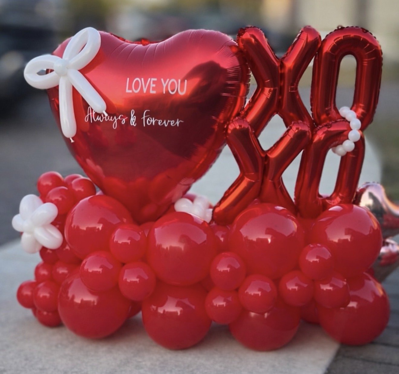 A cluster of red balloons, including a heart-shaped balloon with written message and the word 'XOXO' in balloons, decorated with white balloon flowers, sitting on a surface outdoors.