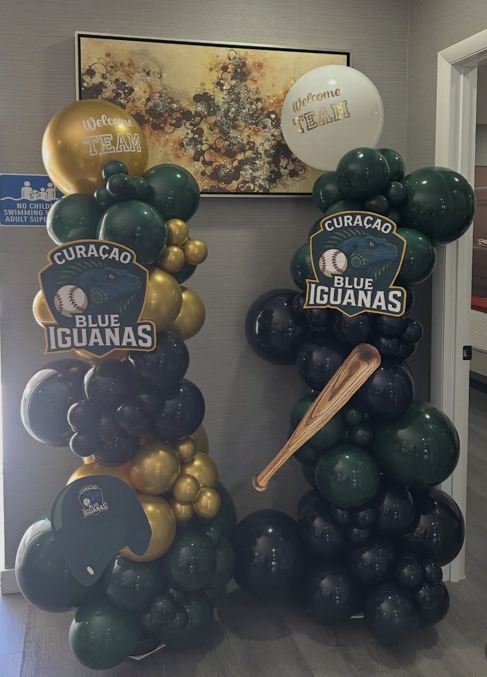 Balloon decorations with logos of Curaçao Blue Iguanas, a baseball theme, and welcome team signs, set up in an indoor space.