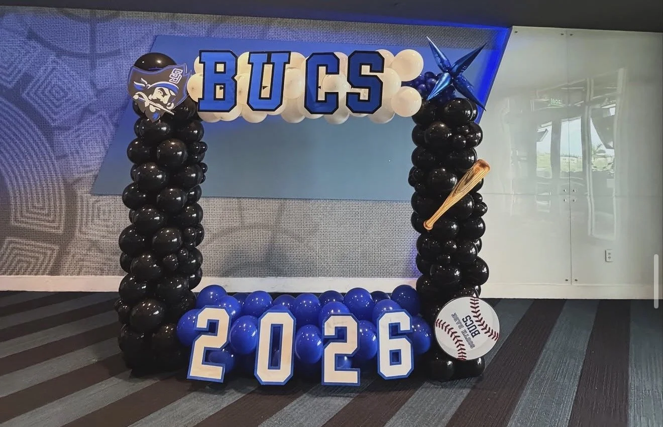 Balloon display with the word 'BUCS' in large letters, decorated with sports-themed items like a helmet, baseball bat, and a baseball, all in blue, black, and white colors. The display includes the year '2026' formed with blue and white block numbers.