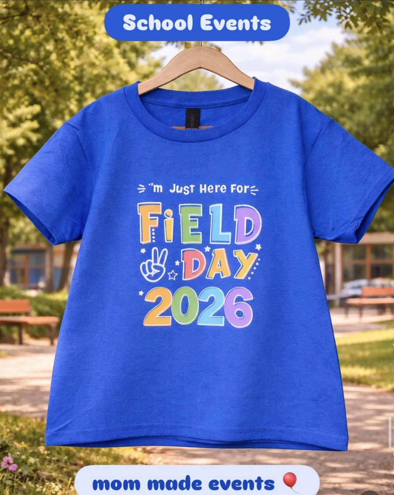 A blue T-shirt hanging on a wooden hanger with colorful text and graphics for a school event in 2026, with a background of trees and a park.