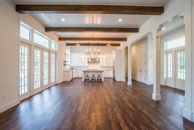 Empty living room with large windows, hardwood floors, white walls, and exposed ceiling beams, open to a kitchen with a central island, chandelier, and barstools.