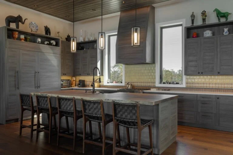 Modern kitchen with gray cabinets, a large island with five chairs, pendant lighting, and decorative animals on the shelves above, with trees visible outside the windows.