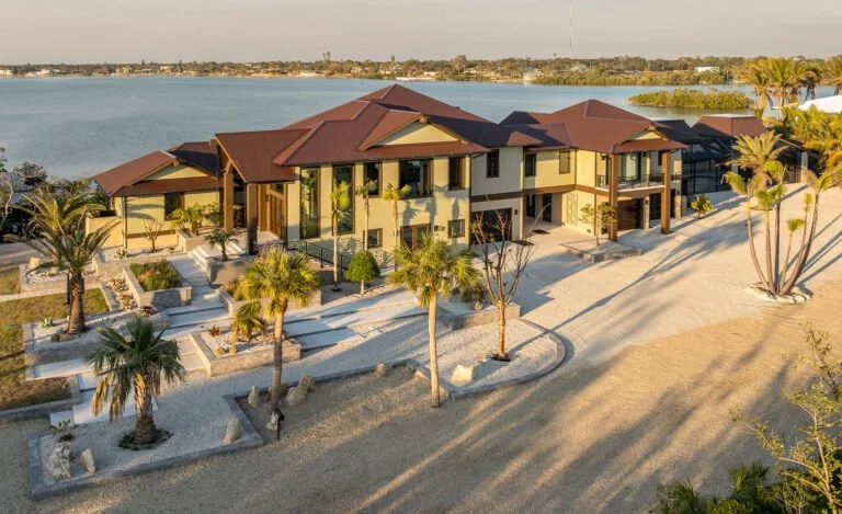 A large modern house with multiple levels, surrounded by palm trees, near a waterfront with a body of water in the background.