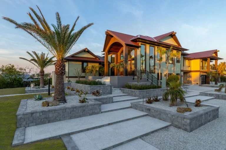 Modern house with large glass windows, surrounded by palm trees and landscaped yard with concrete steps and gravel paths.
