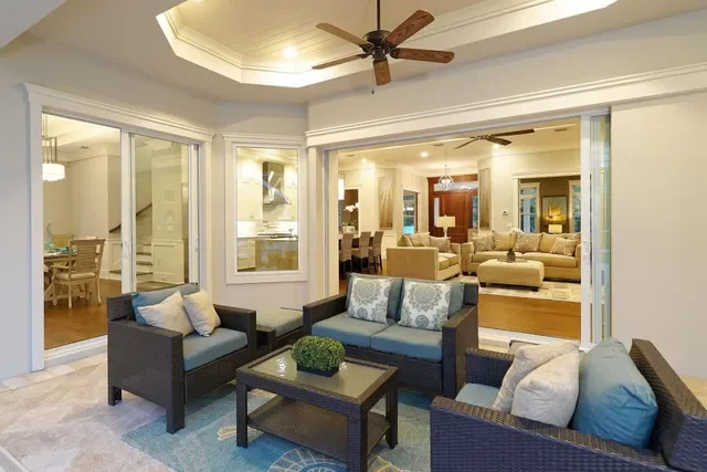 A spacious, well-lit living room with wicker furniture, including a sofa and armchairs with cushions, a coffee table with a small plant, and large sliding glass doors leading to other rooms.