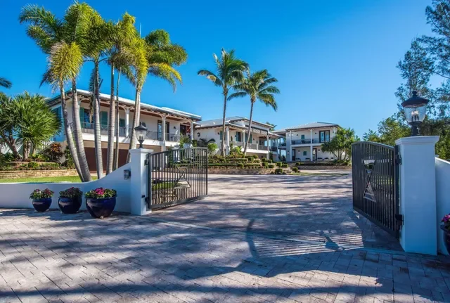 Gated entrance to a large residence with palm trees, a paved driveway, and modern white townhouses in the background.