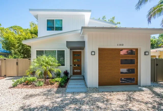 Modern two-story house with white exterior, wooden front door, and wooden garage door, surrounded by landscaping with palm trees and gravel driveway.