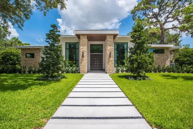 Modern house with a stone facade, large windows, small trees, and a concrete pathway leading to the front door, surrounded by a well-kept lawn and tall trees under a partly cloudy sky.