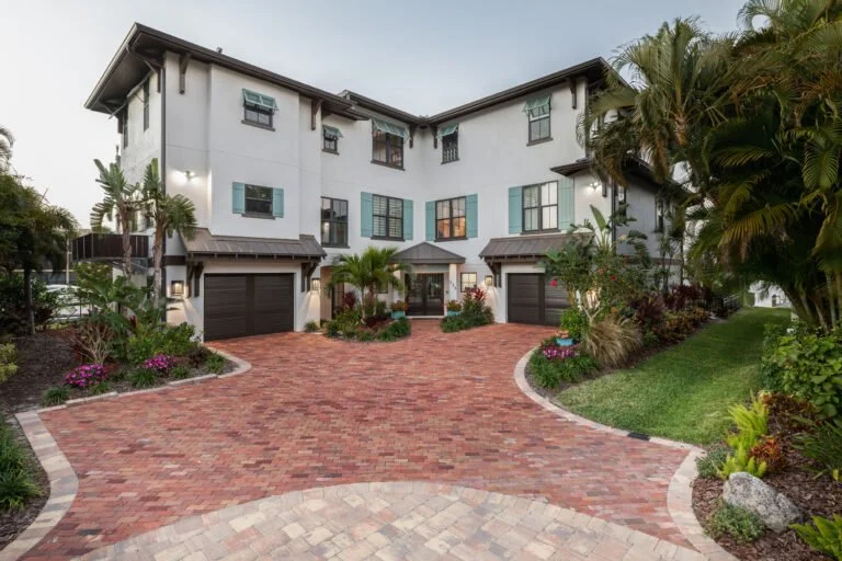A large, multi-story white house with a curved red brick driveway and lush tropical landscaping, including palm trees and colorful plants.
