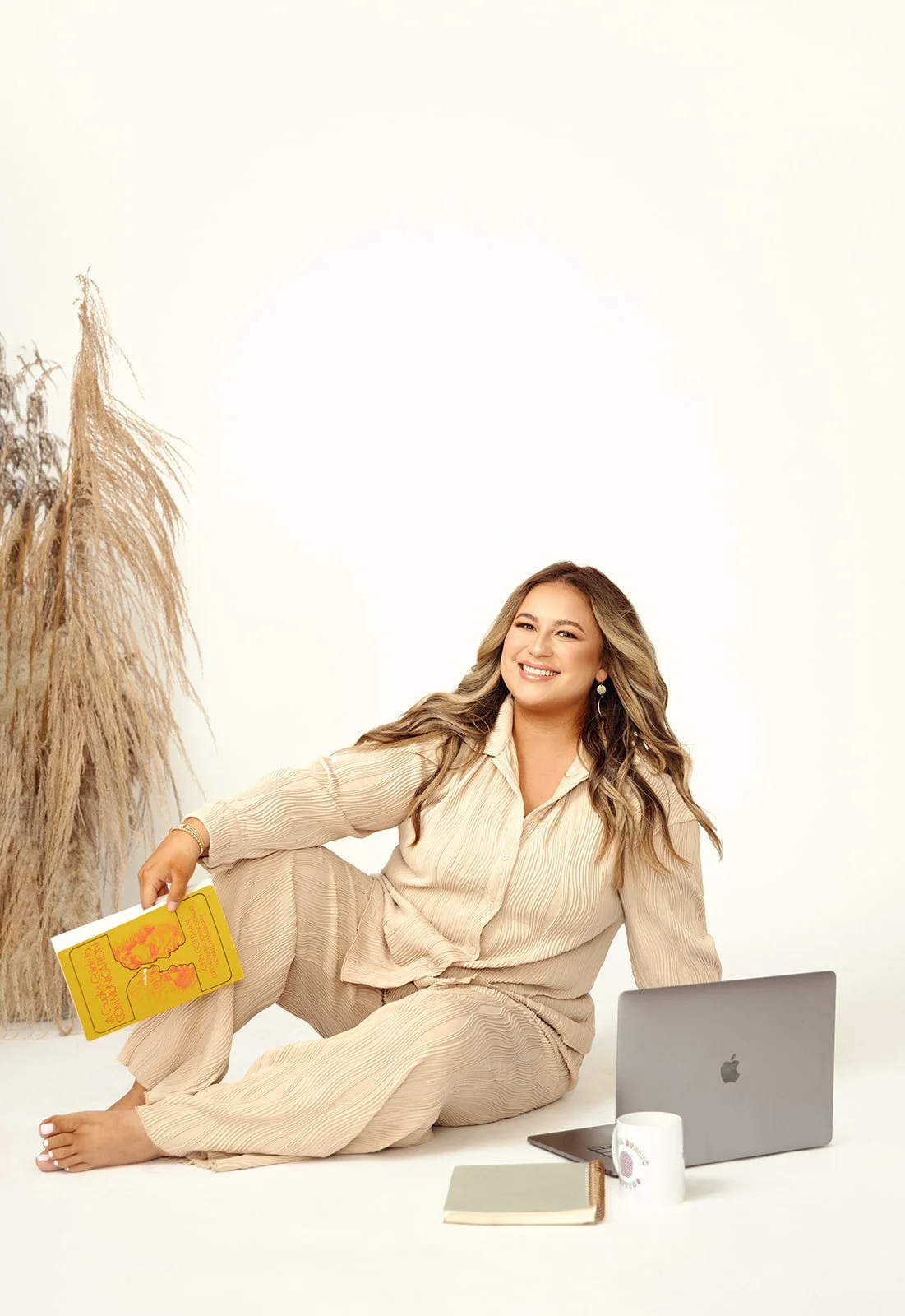 Smiling woman sitting on the floor with a laptop, holding a yellow book, with a notebook and mug nearby, on a white background.