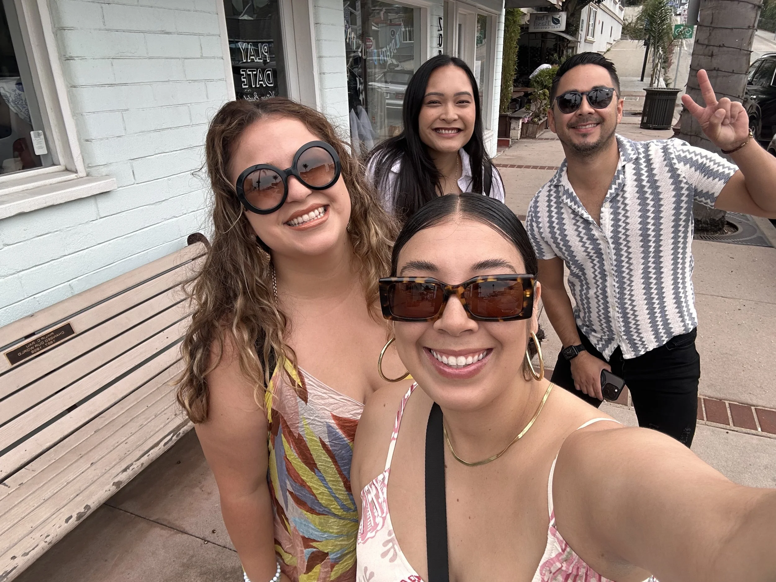 A group of four friends taking a selfie outside a restaurant, all smiling and wearing sunglasses, with a white brick building and sidewalk in the background.