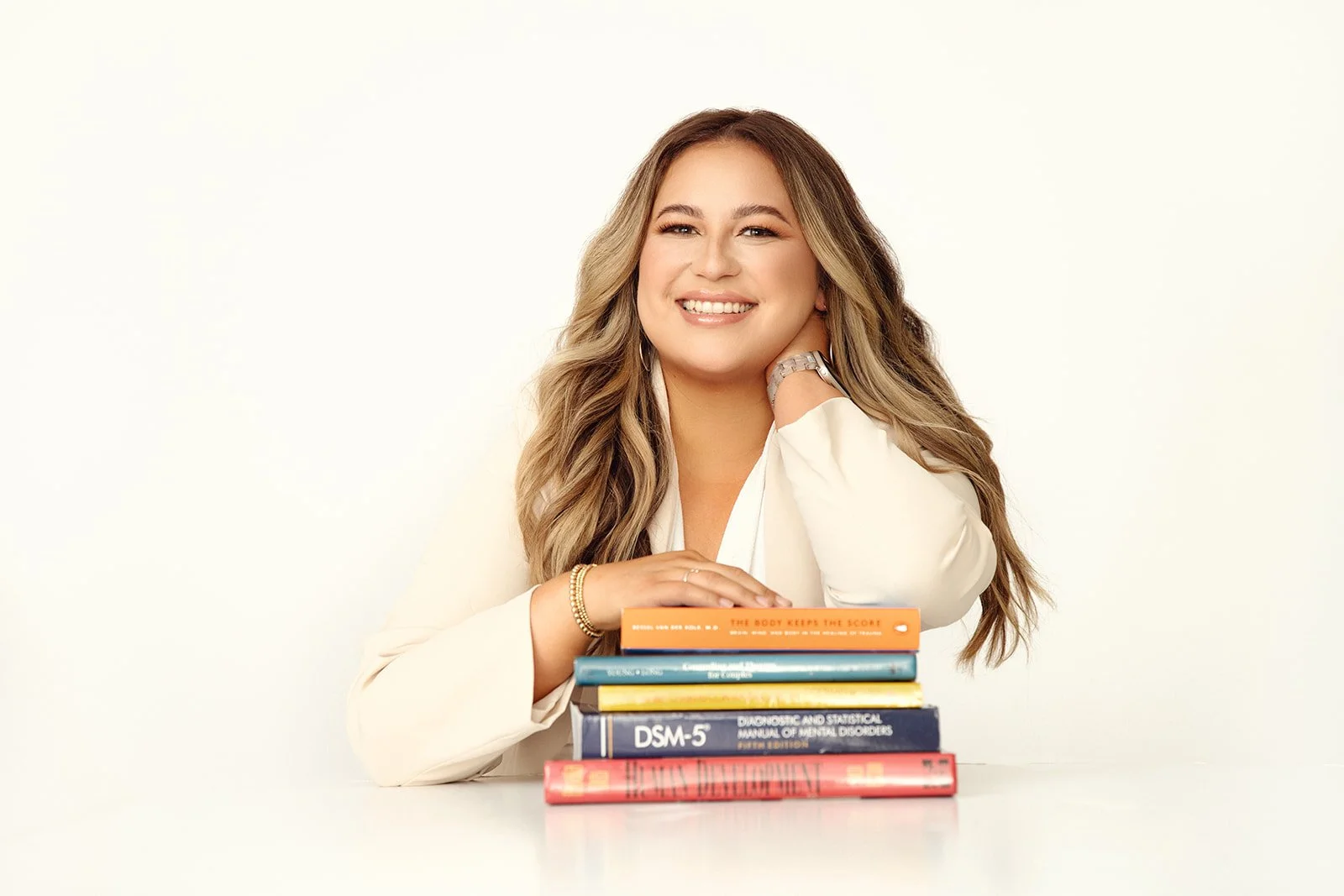 A woman with long wavy hair smiling at the camera, resting her chin on her left hand, with a stack of books on her right side, against a plain white background.