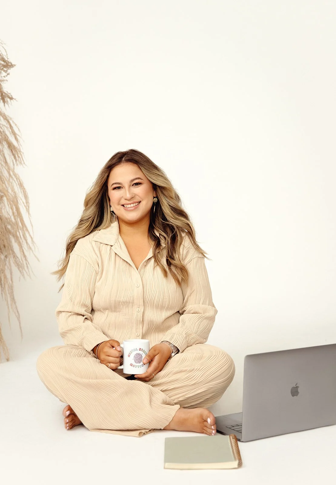 A woman sitting cross-legged on the floor, smiling, holding a coffee mug, with a laptop and notebook in front of her, against a plain white background.