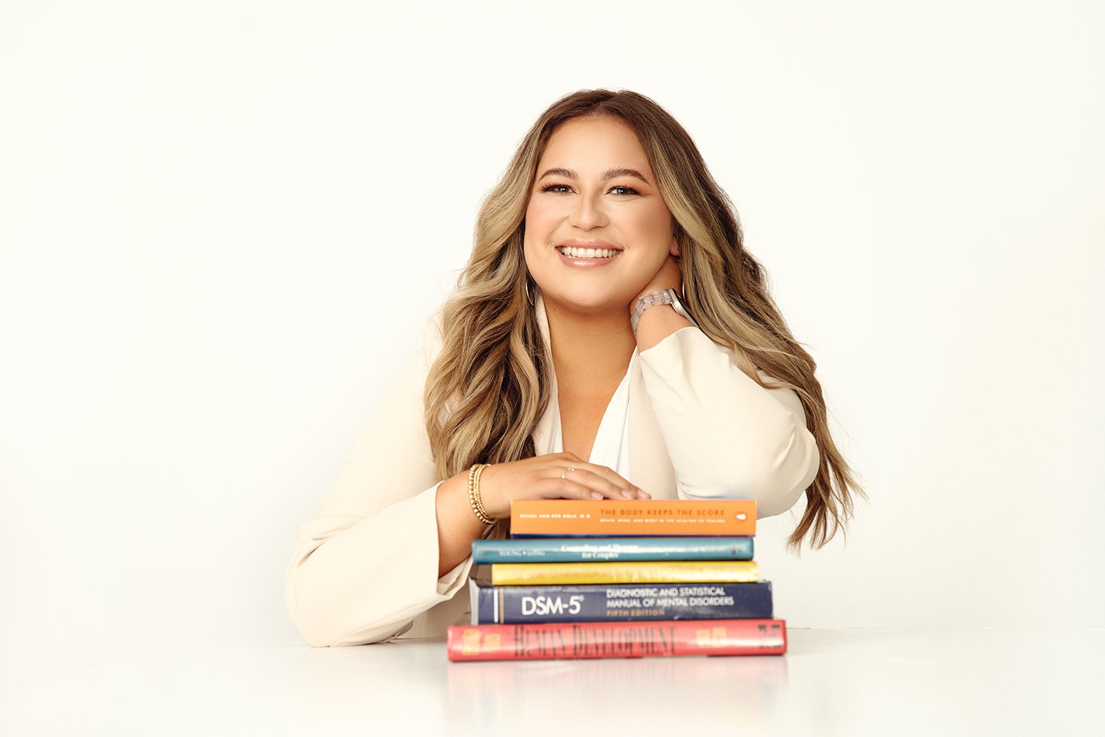 A smiling woman with long wavy hair sits at a white table with a stack of books in front of her, including medical and psychological titles.