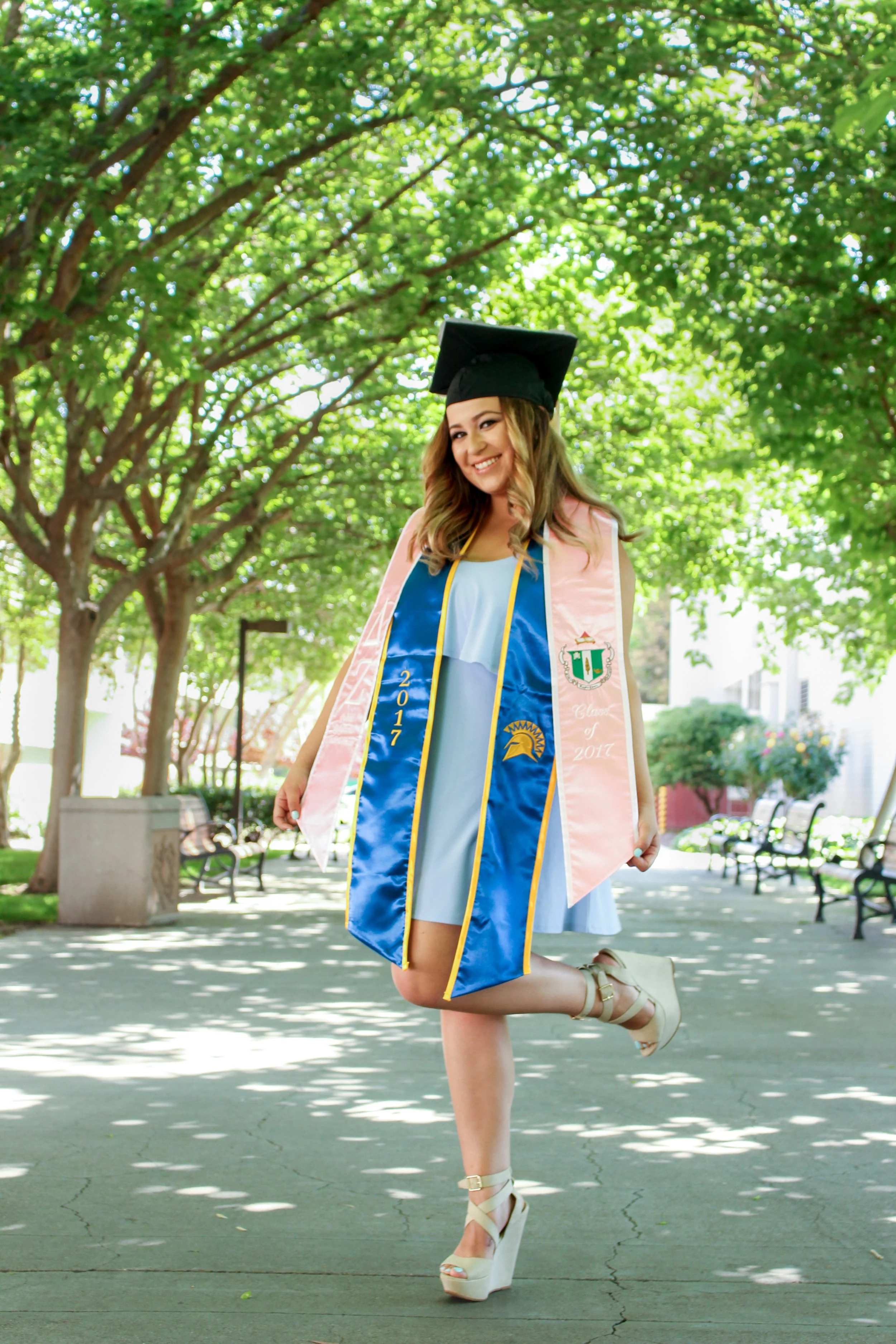 A young woman in a graduation cap and gown is smiling and kneeling outdoors under a tree with green leaves. She is wearing a pink stole and a blue sash with gold accents and a university emblem, and she has beige high-heeled shoes.