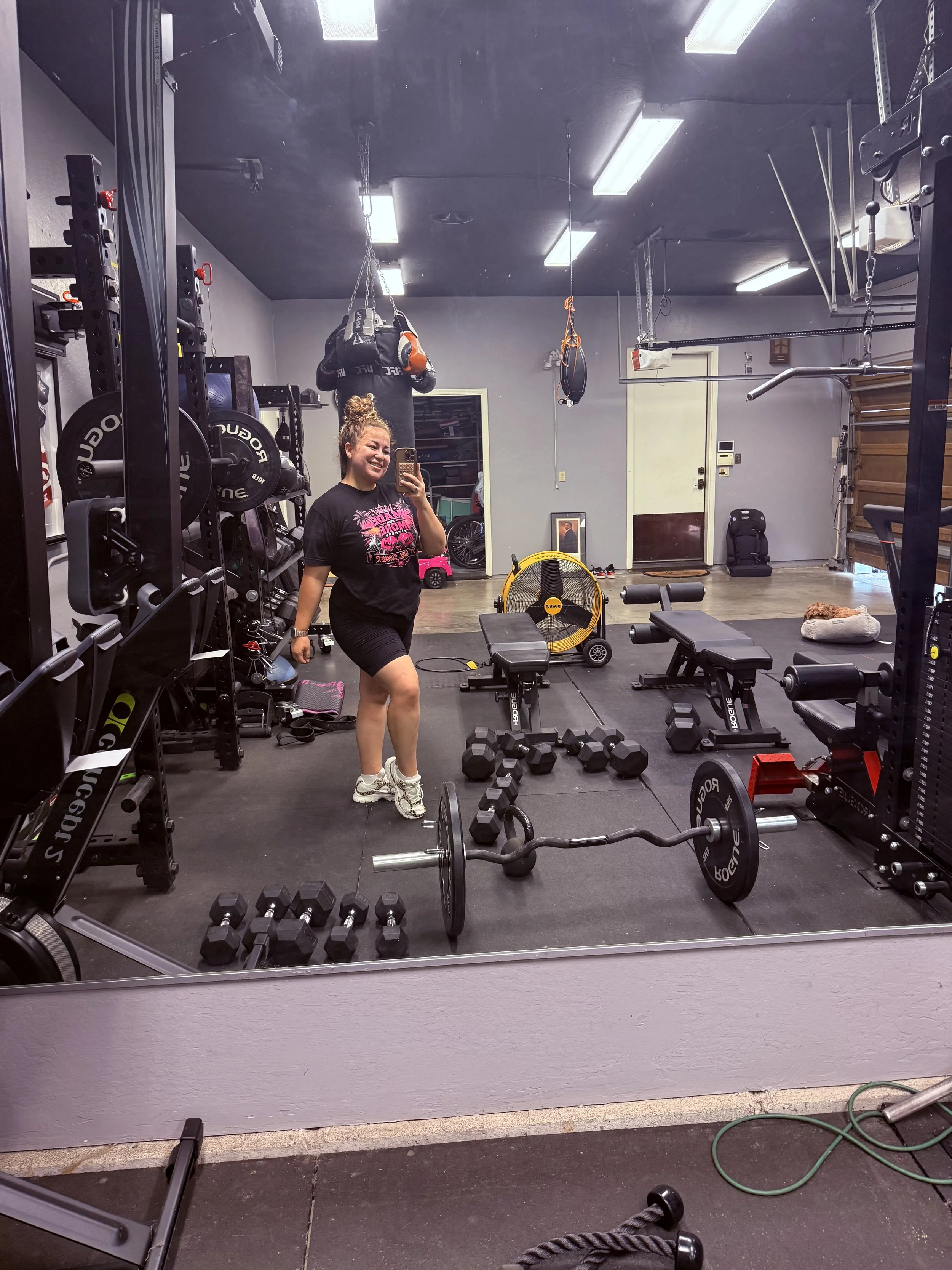 A woman in a black t-shirt and shorts taking a selfie in a gym mirror. The gym has weights, dumbbells, workout bench, and exercise equipment. There is a yellow fan and some children's toys visible in the background.