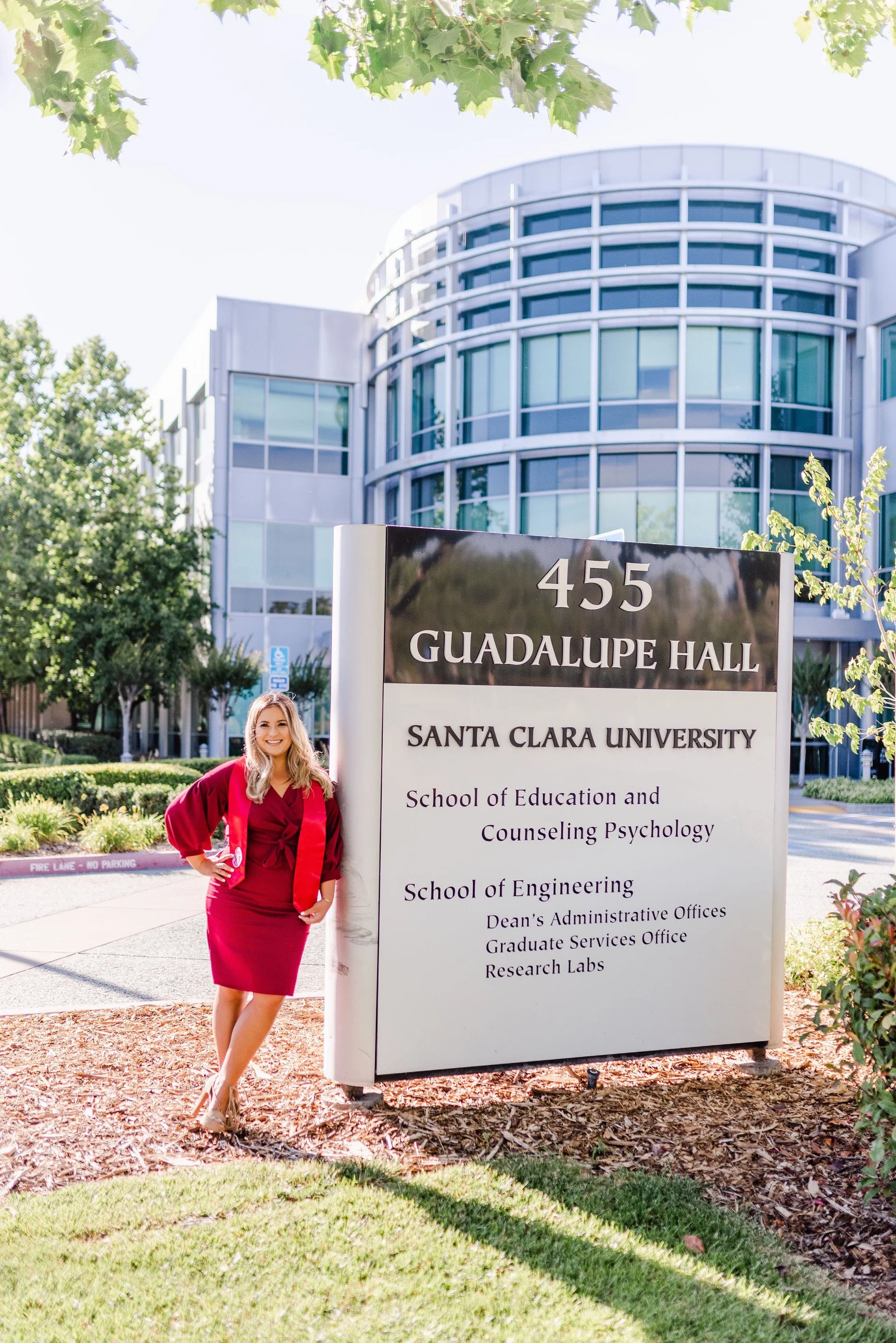 A woman in a red dress standing next to a university sign for Santa Clara University, Guadalupe Hall, smiling outdoors with trees and a modern glass building in the background.
