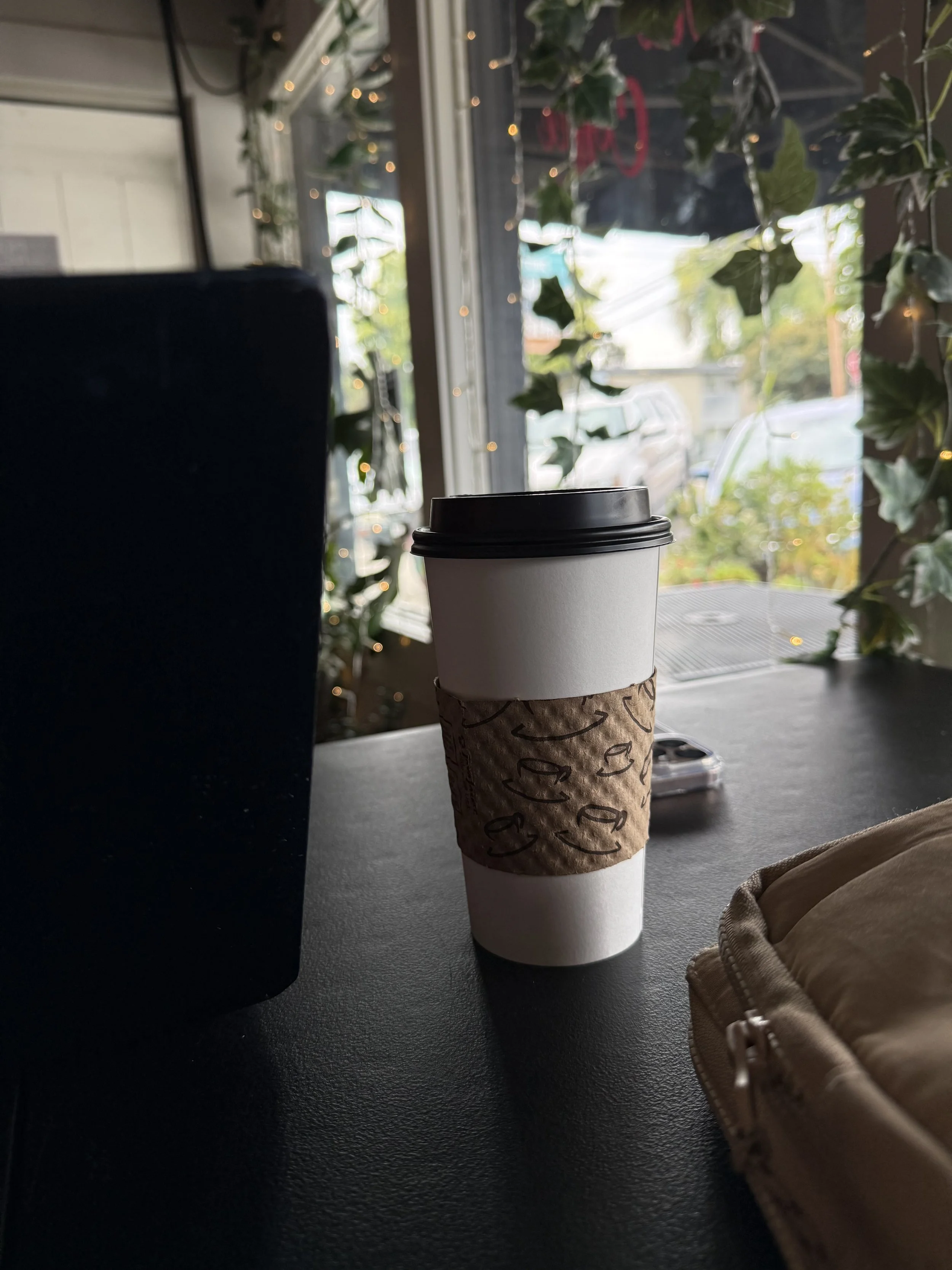 A white disposable coffee cup with a black lid and a brown paper sleeve, placed on a dark table near a beige bag, inside a cafe with a large window and green plants outside.