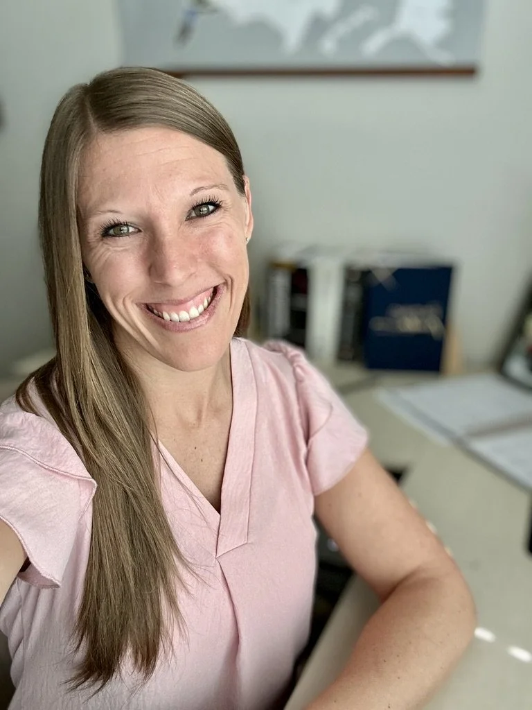 Smiling woman in a pink shirt sitting at a desk in an office with documents and binders in the background.