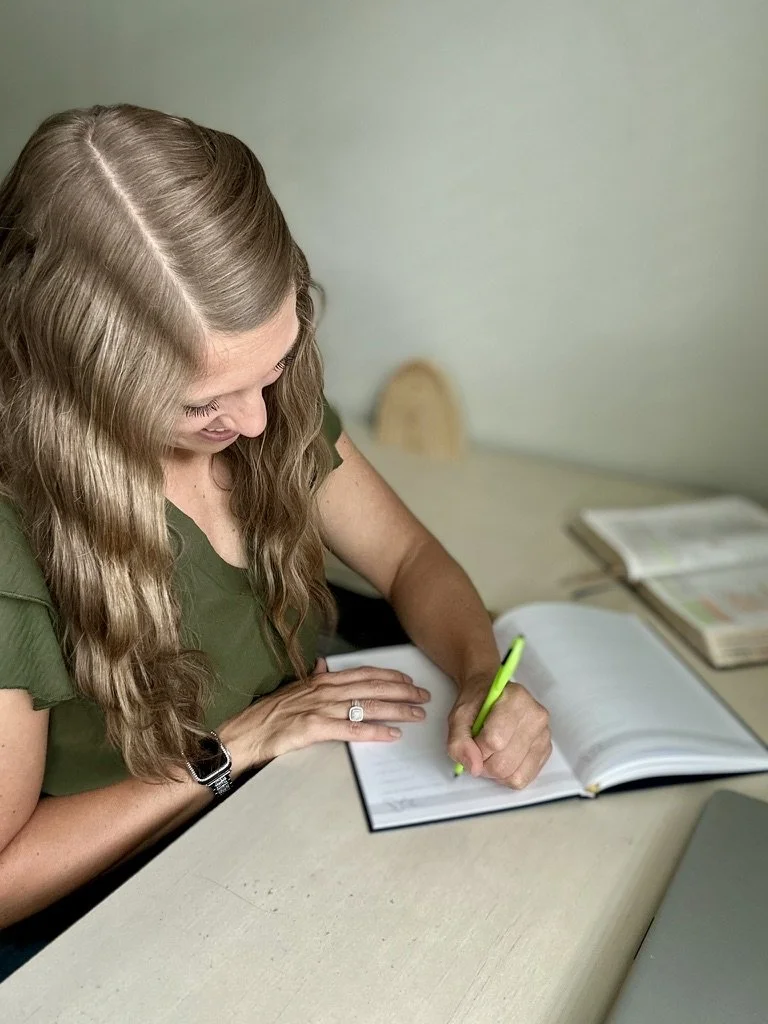 Woman with long wavy hair writing in a notebook at a desk with open books.